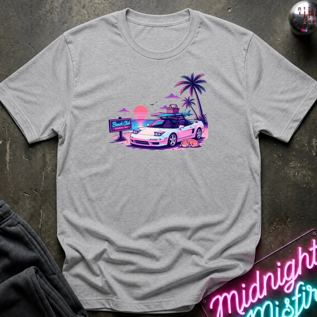 A light heather gray short-sleeve t-shirt laid flat on a dark textured surface, featuring a colorful retro graphic of a white sports car on a beach with palm trees and a sunset.