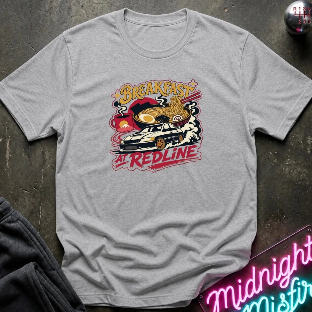 A light gray heather short-sleeve graphic T-shirt laid flat on a dark textured surface. The shirt features a colorful retro-style chest print with the words 'Breakfast at Redline'