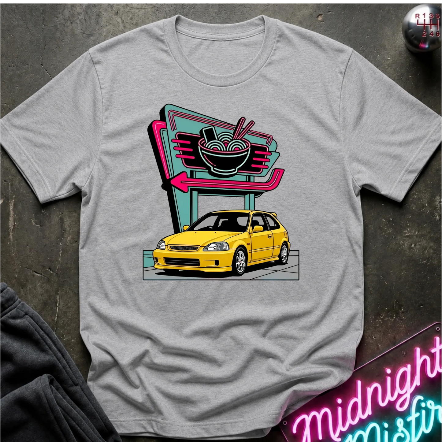 A heather grey  short-sleeve graphic t-shirt laid flat on a dark textured surface. The shirt features a colorful retro-style print of a yellow car parked in front of a neon ramen shop sign.