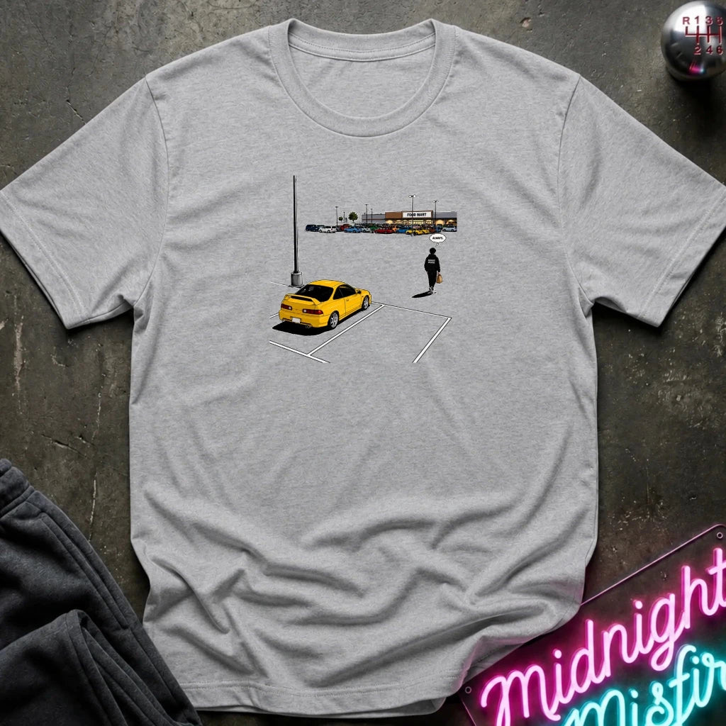 A light heather gray short-sleeve t-shirt laid flat on a dark textured surface, featuring a small graphic print of a yellow car, a parking lot, and a lone figure. 