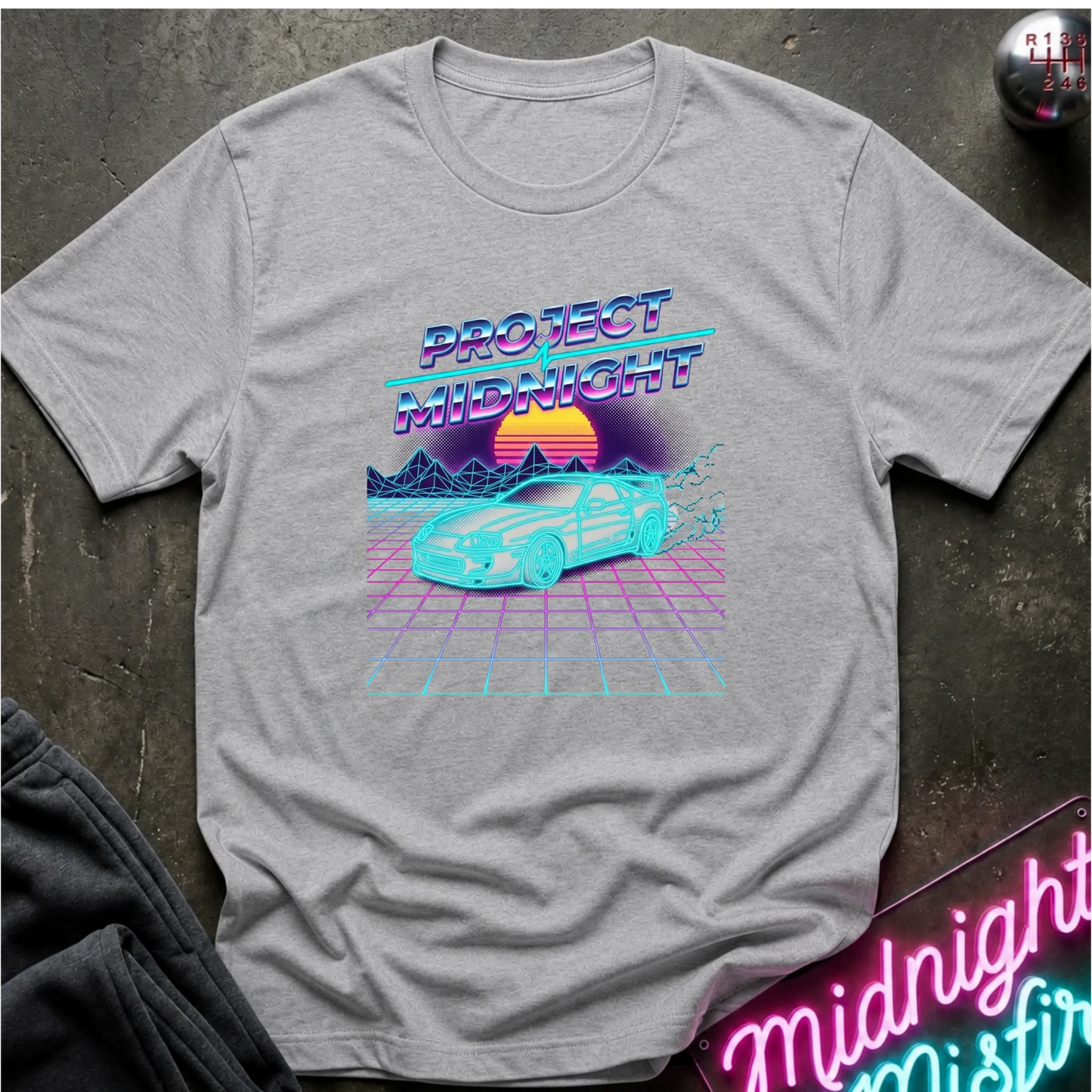 A light gray heather crewneck t-shirt featuring a retro 80s synthwave graphic with the text 'PROJECT MIDNIGHT' and a cyan sports car over a pink grid sunset scene, laid flat on a dark textured surface.