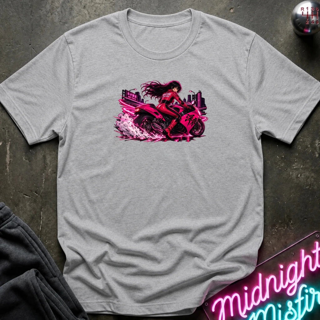 A light heather gray crewneck t-shirt laid flat on a dark textured surface, featuring a vibrant graphic of a woman riding a pink motorcycle with a city skyline in the background. 