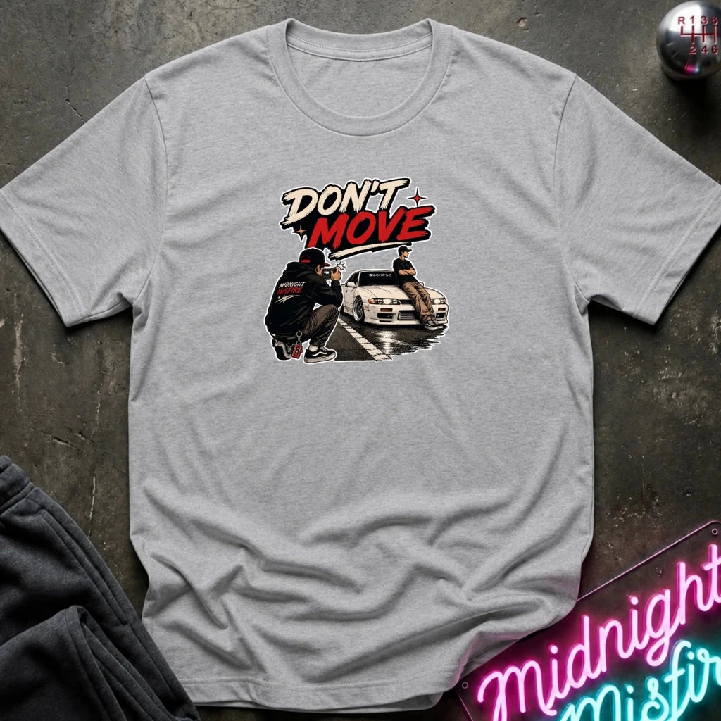 A light gray heather crewneck t-shirt with a graphic print on the front. The graphic features the text 'DON'T MOVE' in stylized red and white lettering above an illustration of a person crouching next to a white sports car.