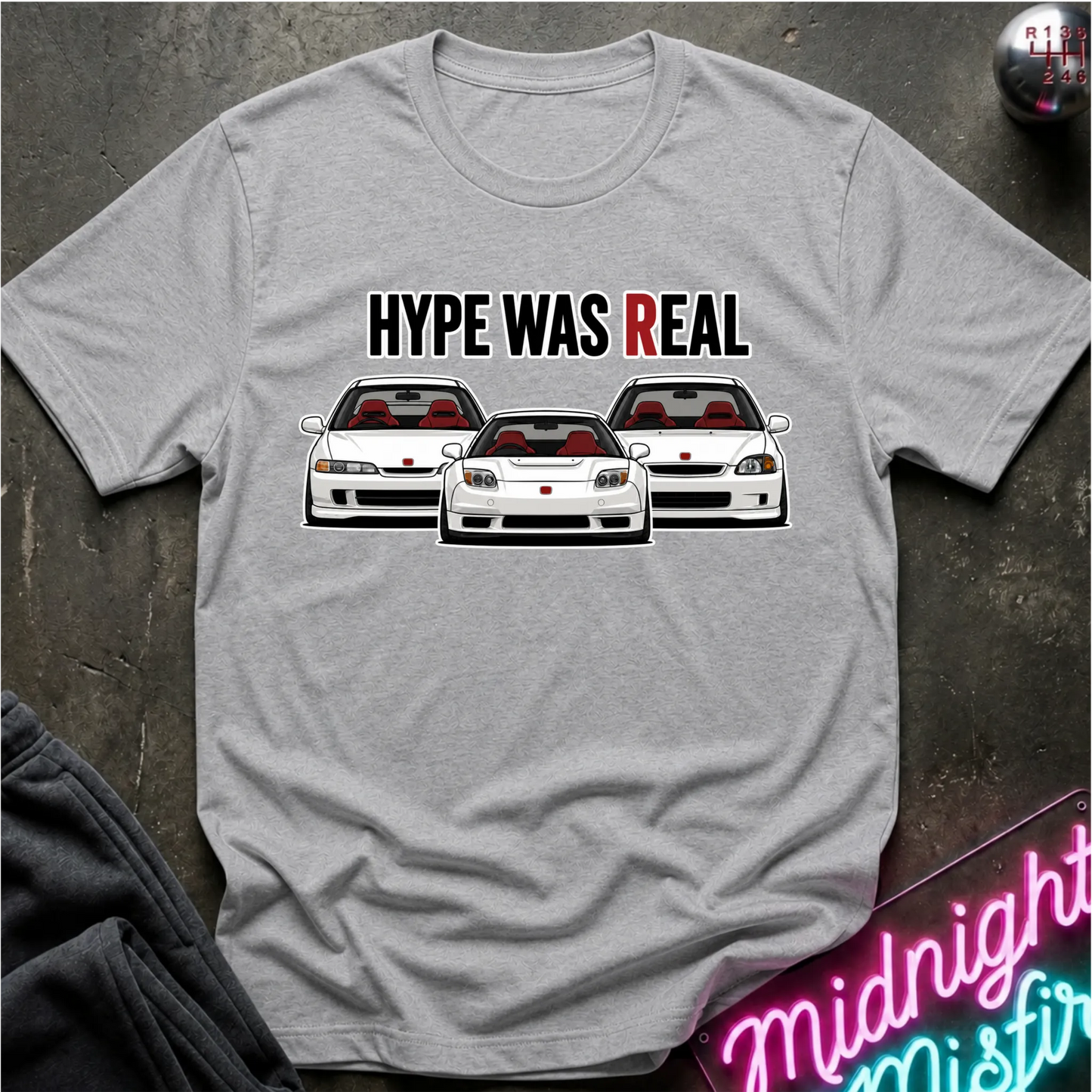 A light gray heather crewneck t-shirt featuring a graphic of three white sports cars with red interiors and the text 'HYPE WAS REAL' in black and red. The shirt is displayed flat on a dark textured surface with a neon sign and dark pants partially visible.