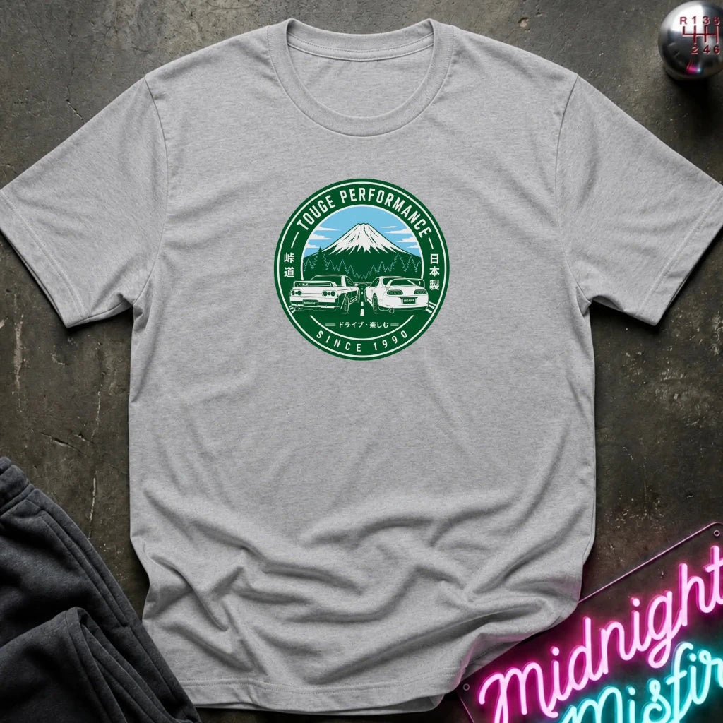 A light gray crewneck t-shirt laid flat on a dark textured surface, featuring a circular green and white graphic print on the chest with a mountain, cars, and the text