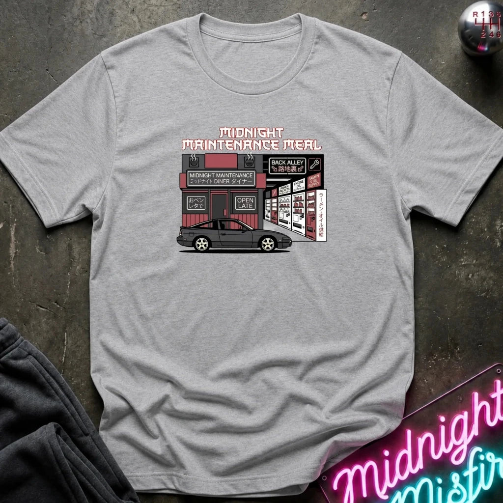 A heather gray short-sleeve t-shirt with a graphic print of a car repair shop and the text 'MIDNIGHT MAINTENANCE MEAL' in pink and black. The shirt is laid flat on a dark textured surface with a neon sign and dark pants partially visible.