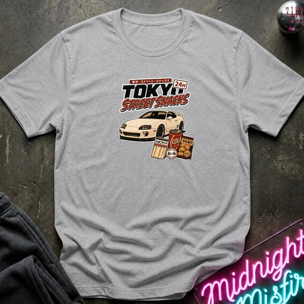 grey short-sleeve graphic T-shirt laid flat on a dark textured surface. The shirt features a central graphic with the text 'Tokyo Street Snacks' above a white sports car and snack illustrations.