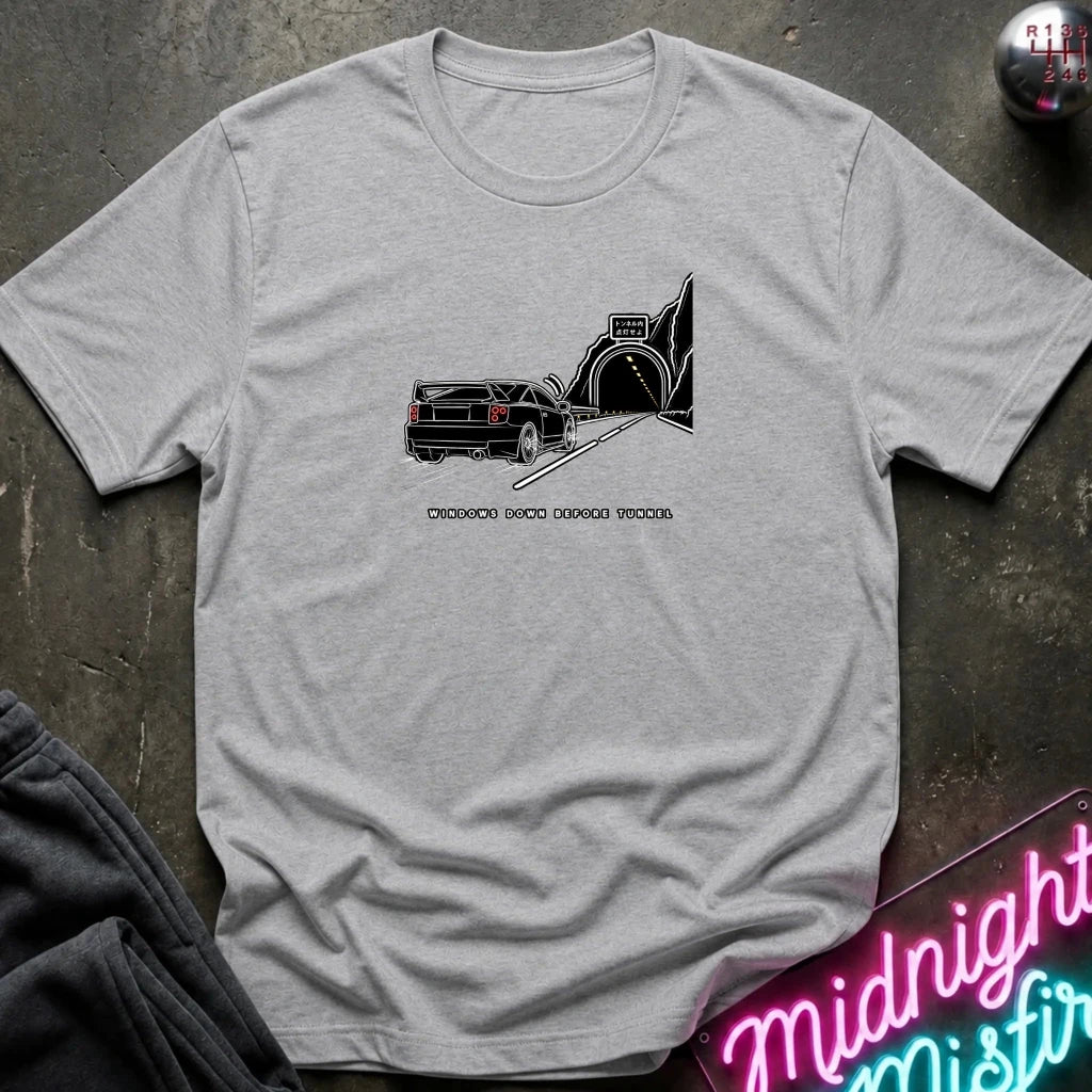 A light gray short-sleeve t-shirt laid flat on a dark textured surface, featuring a black graphic of a classic car in a tunnel with the text.