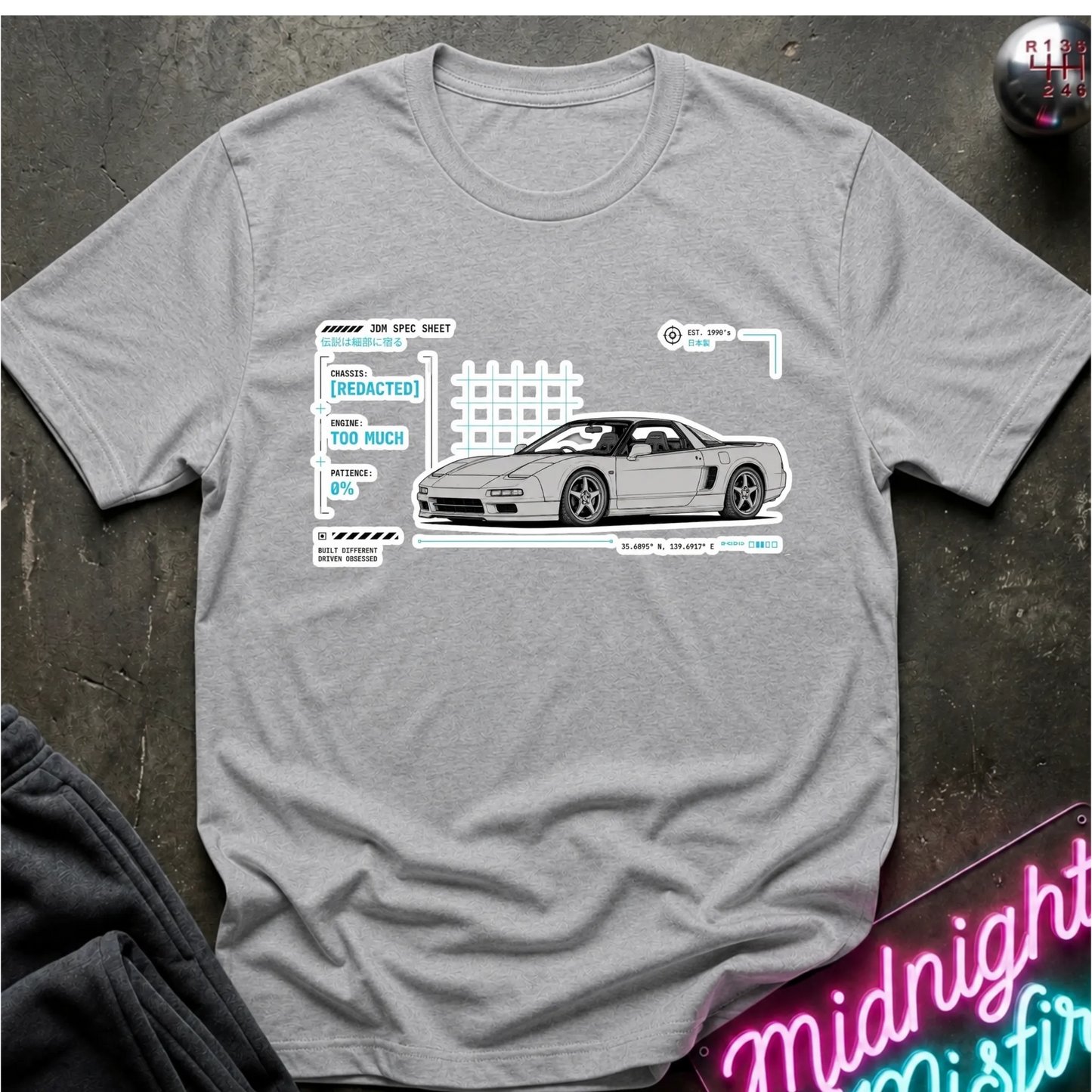 A heather gray short-sleeve t-shirt laid flat on a dark textured surface, featuring a graphic print of a white sports car with technical-style text and line drawings. A neon sign and folded dark fabric are partially visible around the edges.