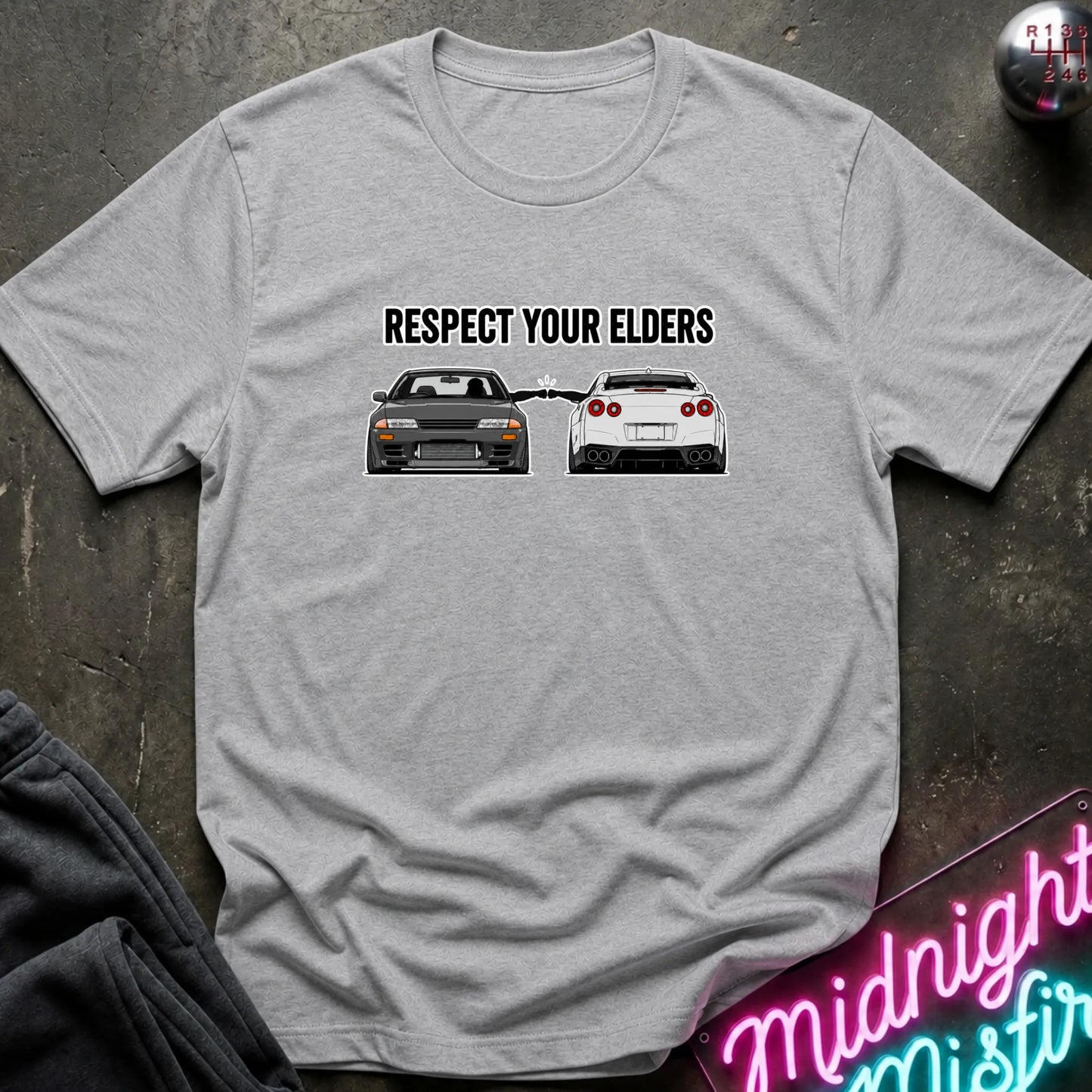 A light gray heather short-sleeve graphic T-shirt laid flat on a dark textured surface. The shirt has a black printed slogan reading 'RESPECT YOUR ELDERS' above two cars, with a neon sign and dark clothing partially visible around it.