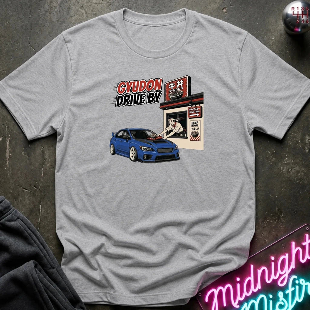 A heather gray short-sleeve graphic t-shirt laid flat on a dark textured surface. The graphic features a blue sports car in front of a Japanese-style shop with the text 'GYUDON DRIVE BY'.