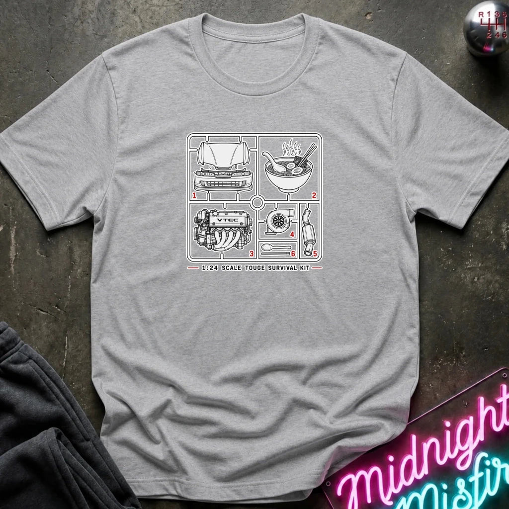 A heather gray short-sleeve t-shirt laid flat on a dark textured surface, featuring a white line-art graphic of a 1:24 scale house survival kit with various tools and ingredients. 