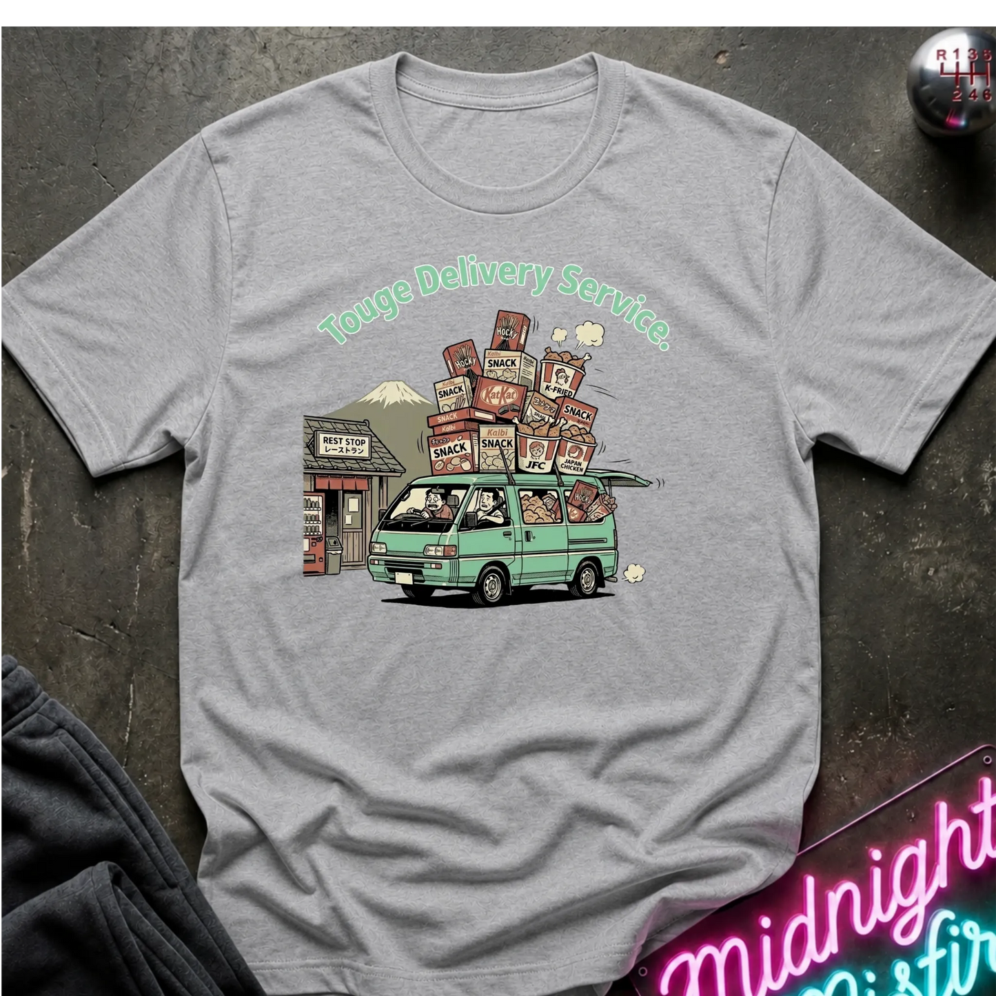 A heather gray short-sleeve t-shirt with a graphic print of a green van carrying stacked cardboard boxes under the text 'Touge Delivery Service.' The shirt is laid flat on a dark textured surface with a neon sign and dark fabric partially visible at the edges.