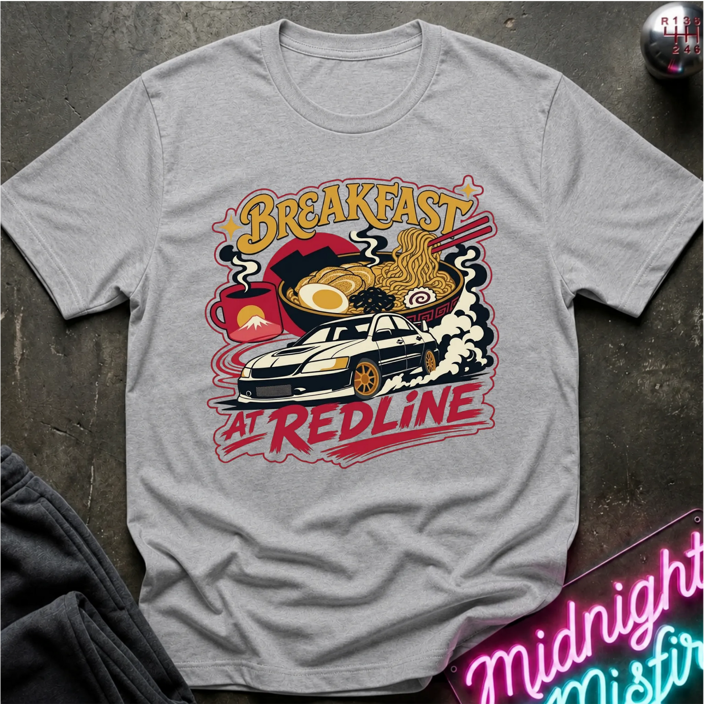 A light gray short-sleeve graphic T-shirt laid flat on a dark textured surface. The shirt features a colorful retro-style print with the words “Breakfast at Redline,” a white car, ramen noodles, an egg, and red abstract shapes.