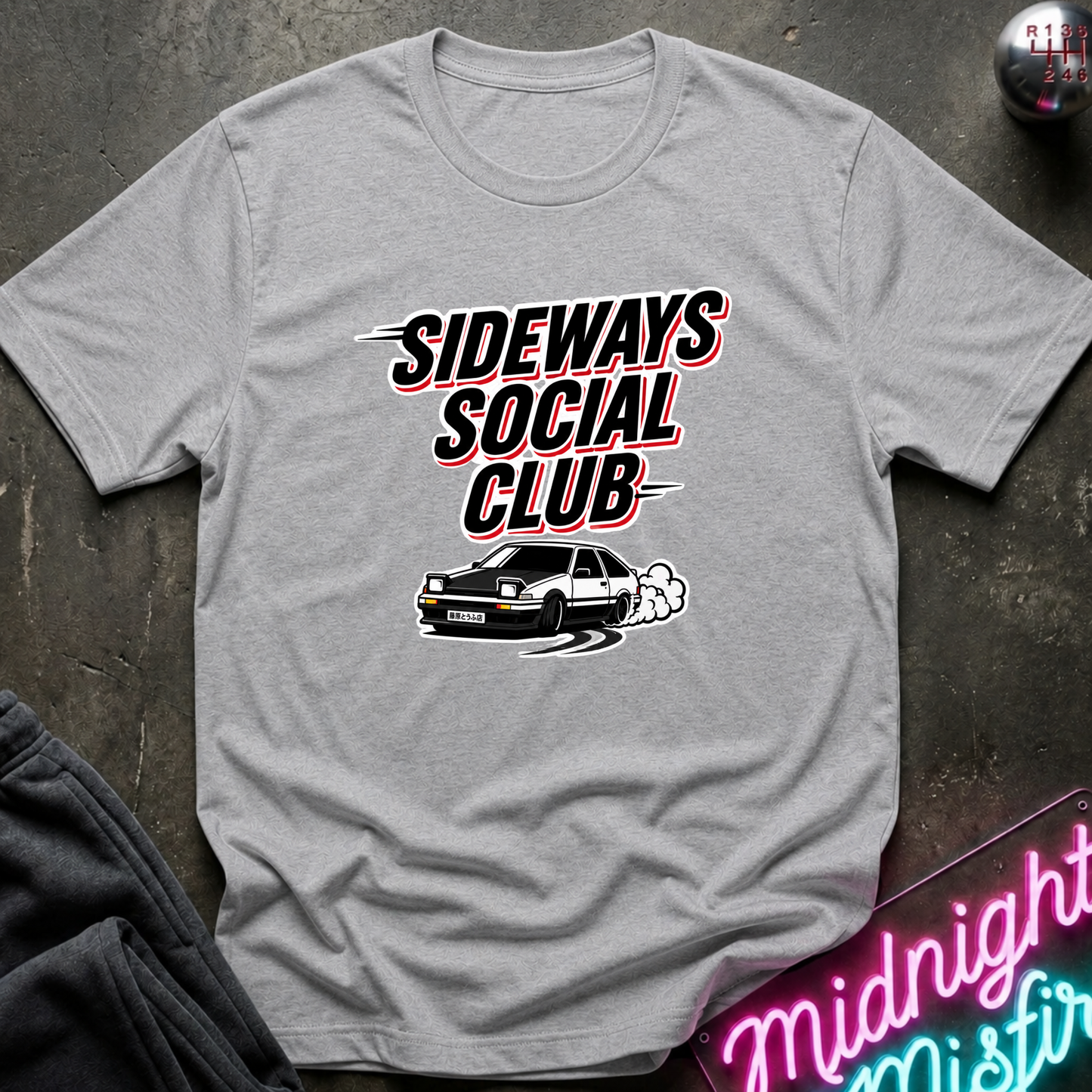 A light gray heather crewneck t-shirt featuring a graphic print of the text 'SIDEWAYS SOCIAL CLUB' in a stylized black, red, and white font, with a black and white car drifting below it. The shirt is laid flat on a dark textured surface with a neon sign and dark pants partially visible.