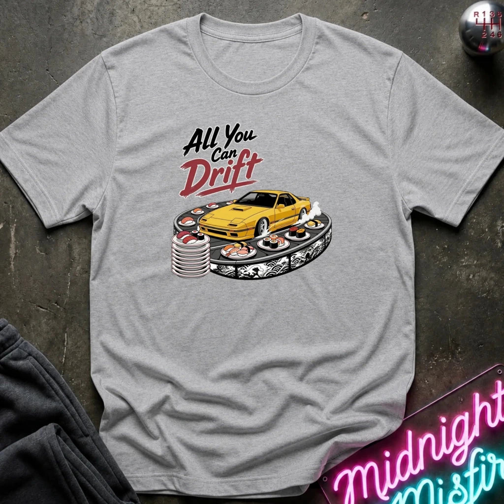 A light gray crewneck t-shirt featuring a graphic of a yellow sports car drifting on a stack of tires, with the text 'All You Can Drift' in black and red. The shirt is displayed flat on a dark textured surface with a neon sign and dark pants partially visible.