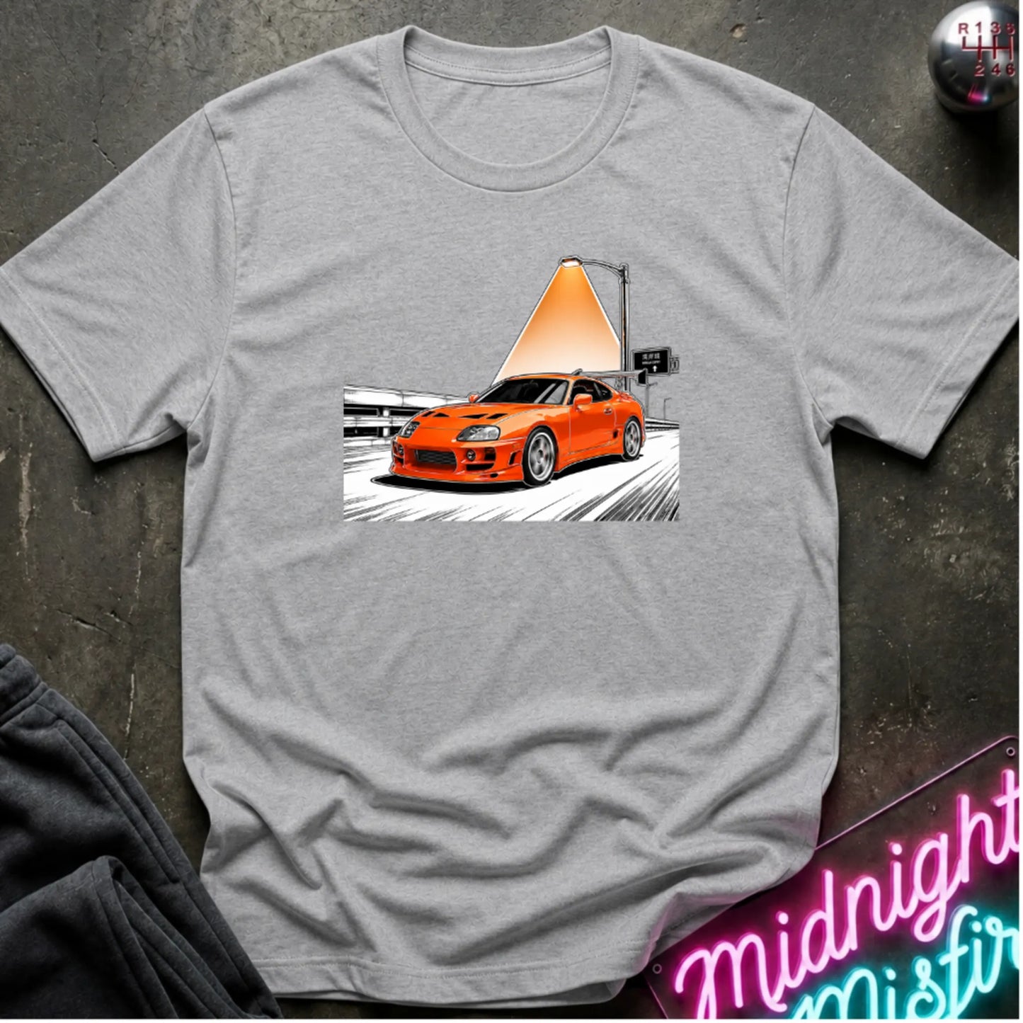 A heather gray short-sleeve t-shirt laid flat on a dark textured surface, featuring a graphic print of an orange sports car speeding on highway.