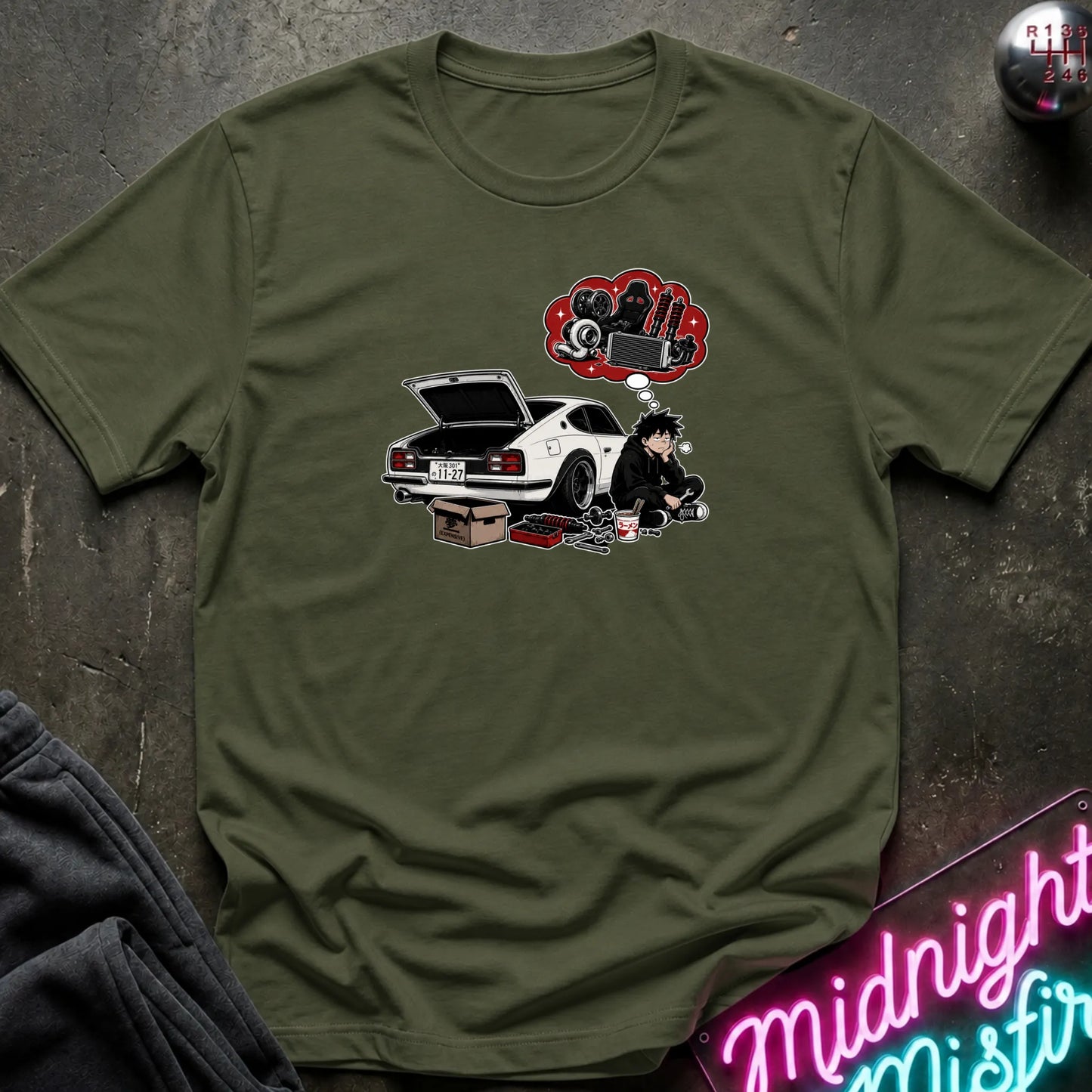 An olive green short-sleeve graphic t-shirt laid flat on a dark textured surface. The graphic features a white sports car with its trunk open, a cartoon character sitting beside it, and a red thought bubble filled with car parts.