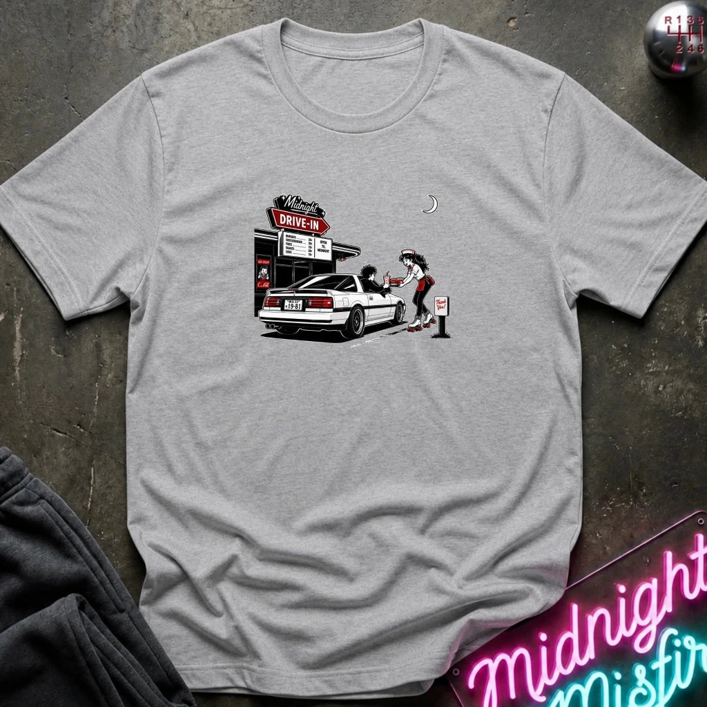 A heather gray short-sleeve t-shirt laid flat on a dark textured surface, featuring a graphic print of a woman pumping gas at a drive-in station next to a white car. 
