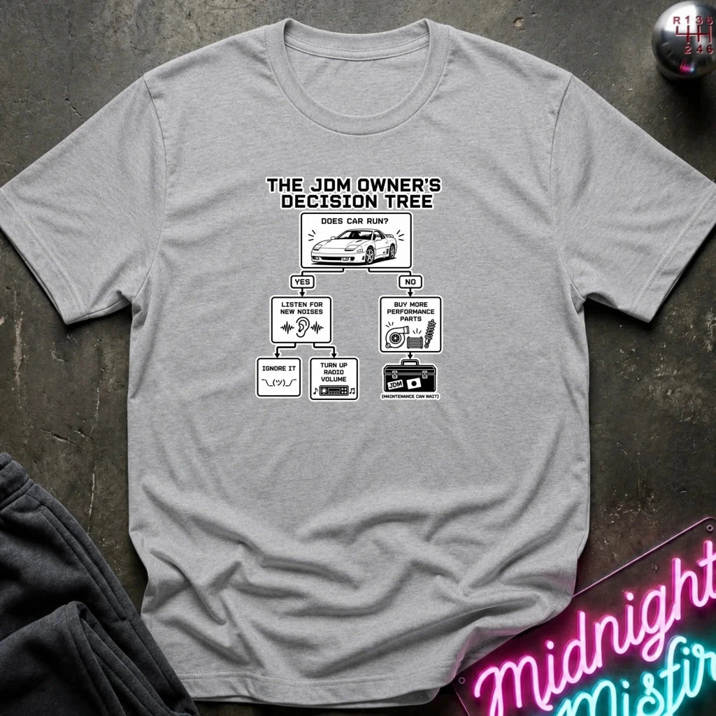 A light gray crewneck t-shirt with a black and white graphic print titled 'THE JDM OWNER'S DECISION TREE' showing a car and various driving-related diagrams. The shirt is laid flat on a dark textured surface with a neon sign and dark pants partially visible in the corners.