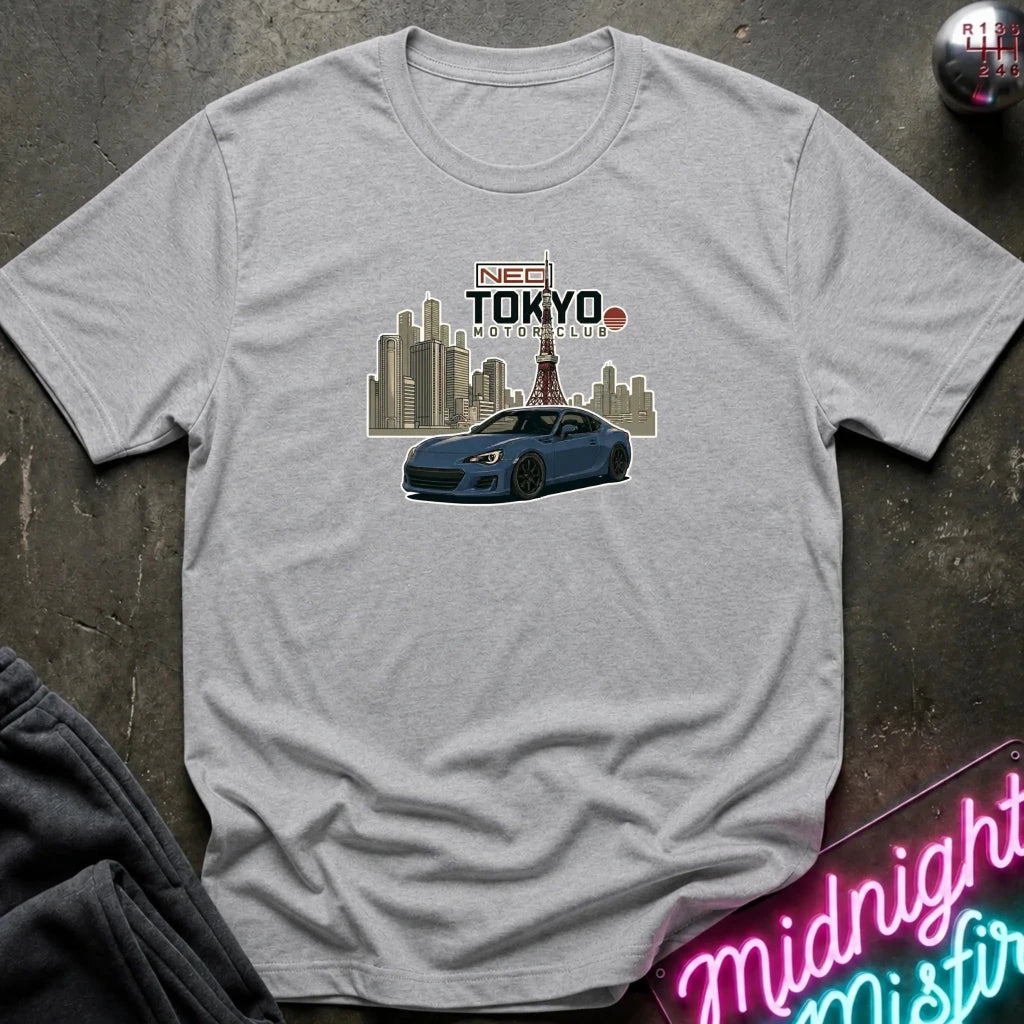 A light gray crewneck t-shirt laid flat on a dark textured surface, featuring a graphic print of a blue sports car in front of a Tokyo city skyline with the text 'NEED TOKYO MOTORS CLUB'.