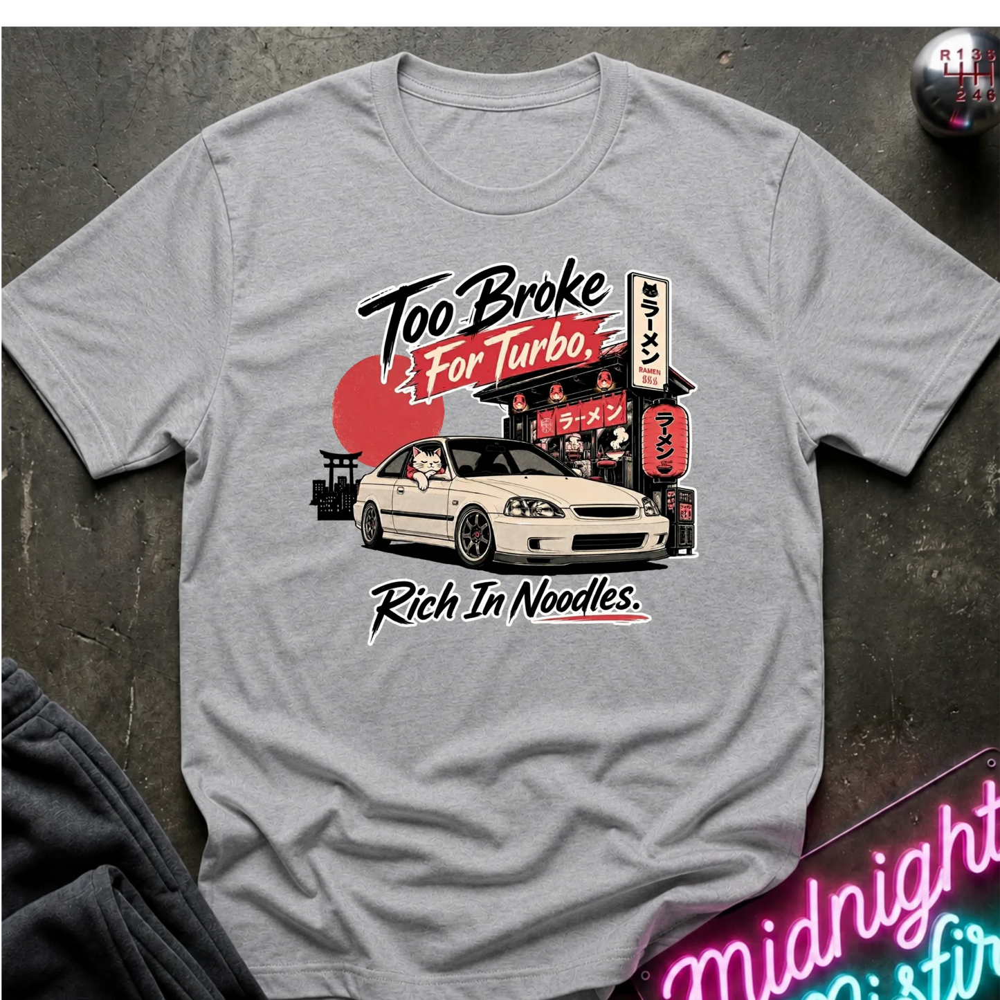 Gray  short-sleeve graphic T-shirt laid flat on a dark textured surface. The shirt features a printed design of a white sports car in front of a Japanese-style street scene with a red sun and neon signs, plus the text 'Too Broke For Turbo' and 'Rich In Noodles.