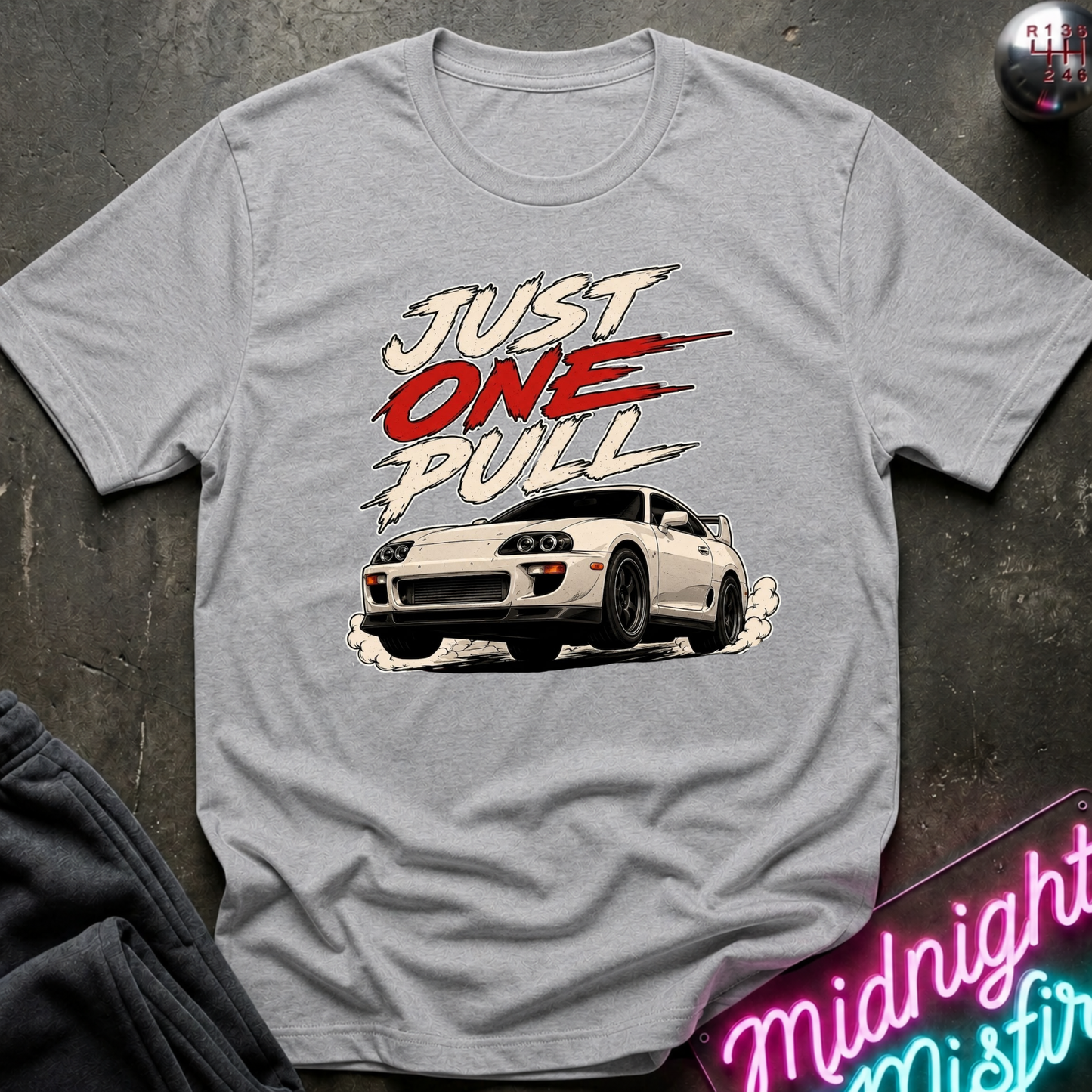 A heather gray crewneck t-shirt featuring a graphic print of a white sports car and the text 'JUST ONE PULL' in white and red, laid flat on a dark textured surface.