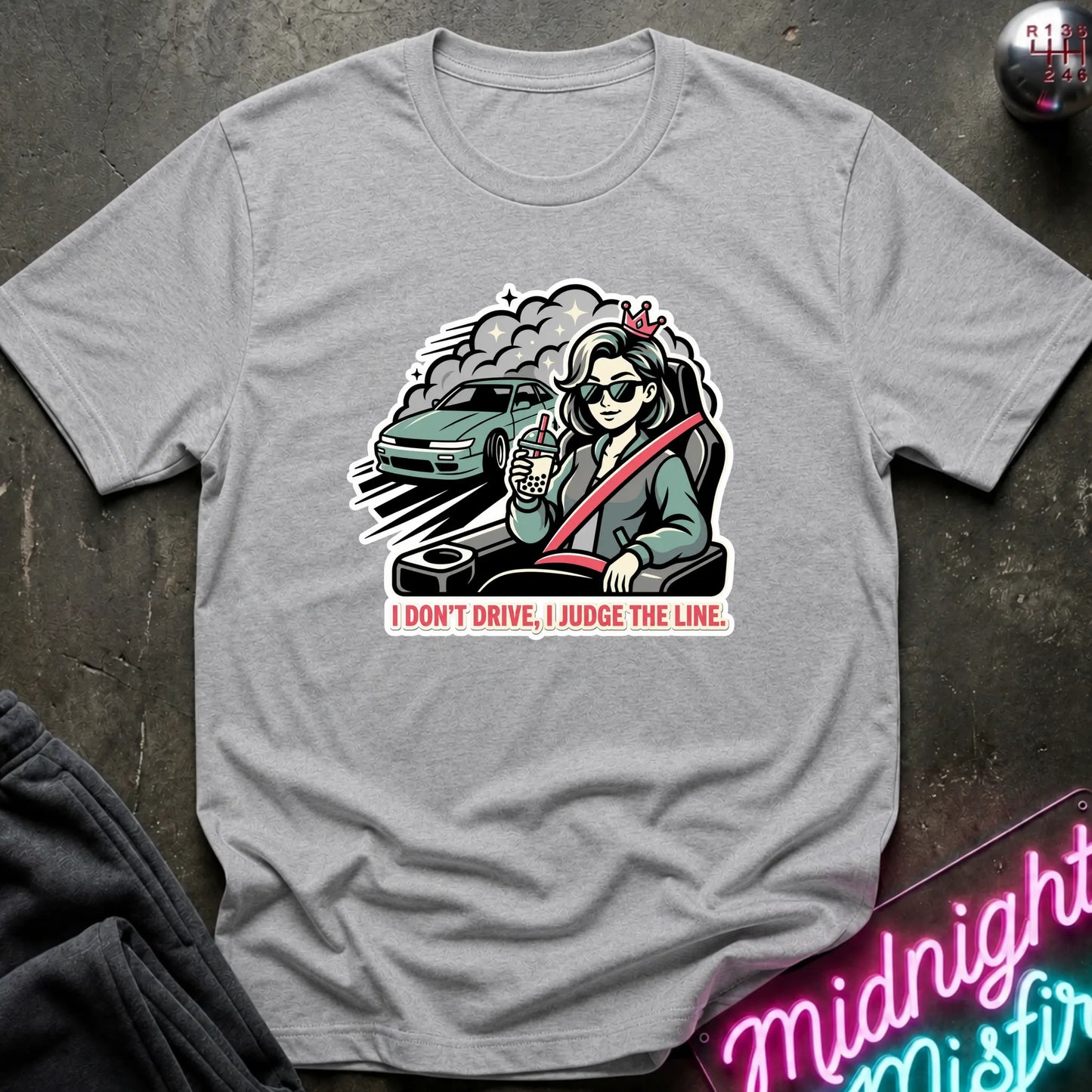 A heather gray short-sleeve t-shirt laid flat on a dark textured surface, featuring a graphic of a woman in sunglasses driving a green car with the text 'I DON'T DRIVE, I JUDGE THE LINE.'.