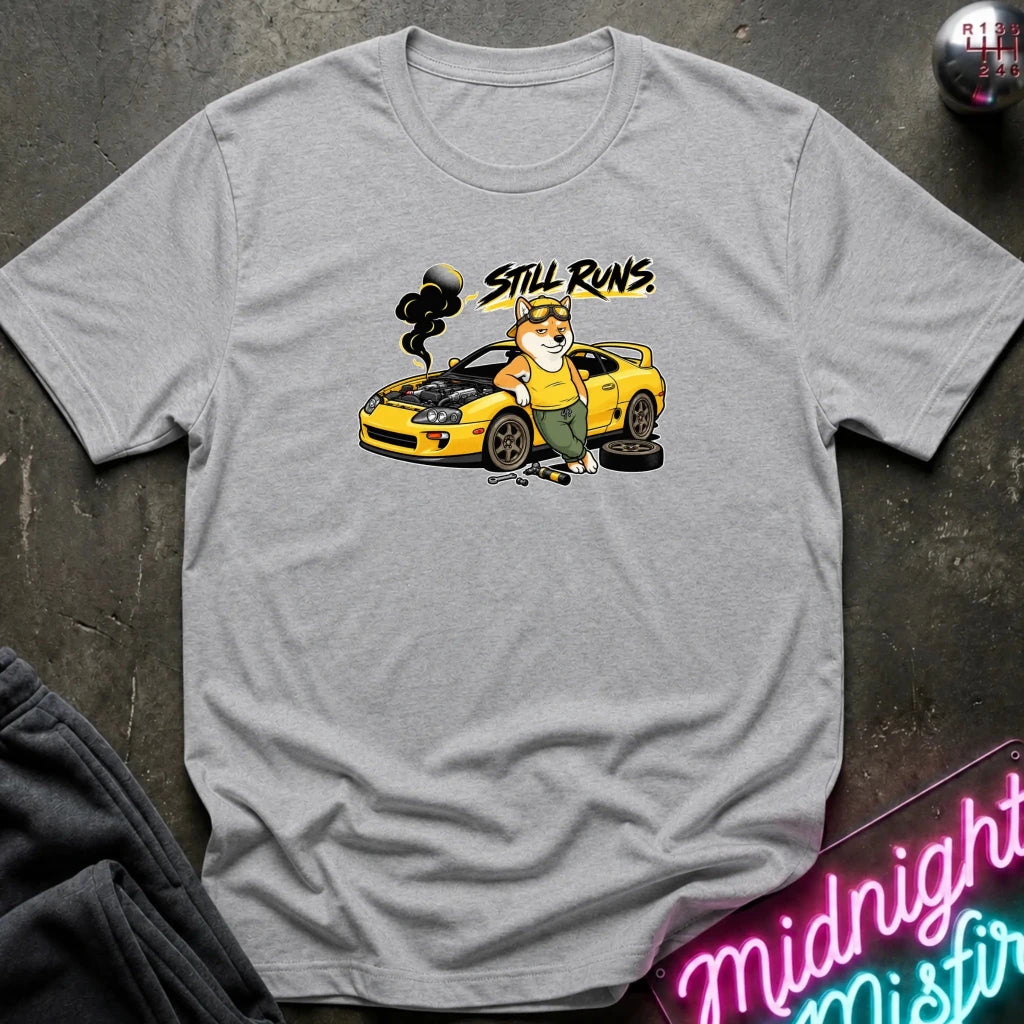 A heather gray short-sleeve t-shirt with a graphic print of a cartoon dog in green overalls working on a yellow sports car, with the text 'STILL RUNS.' above it.