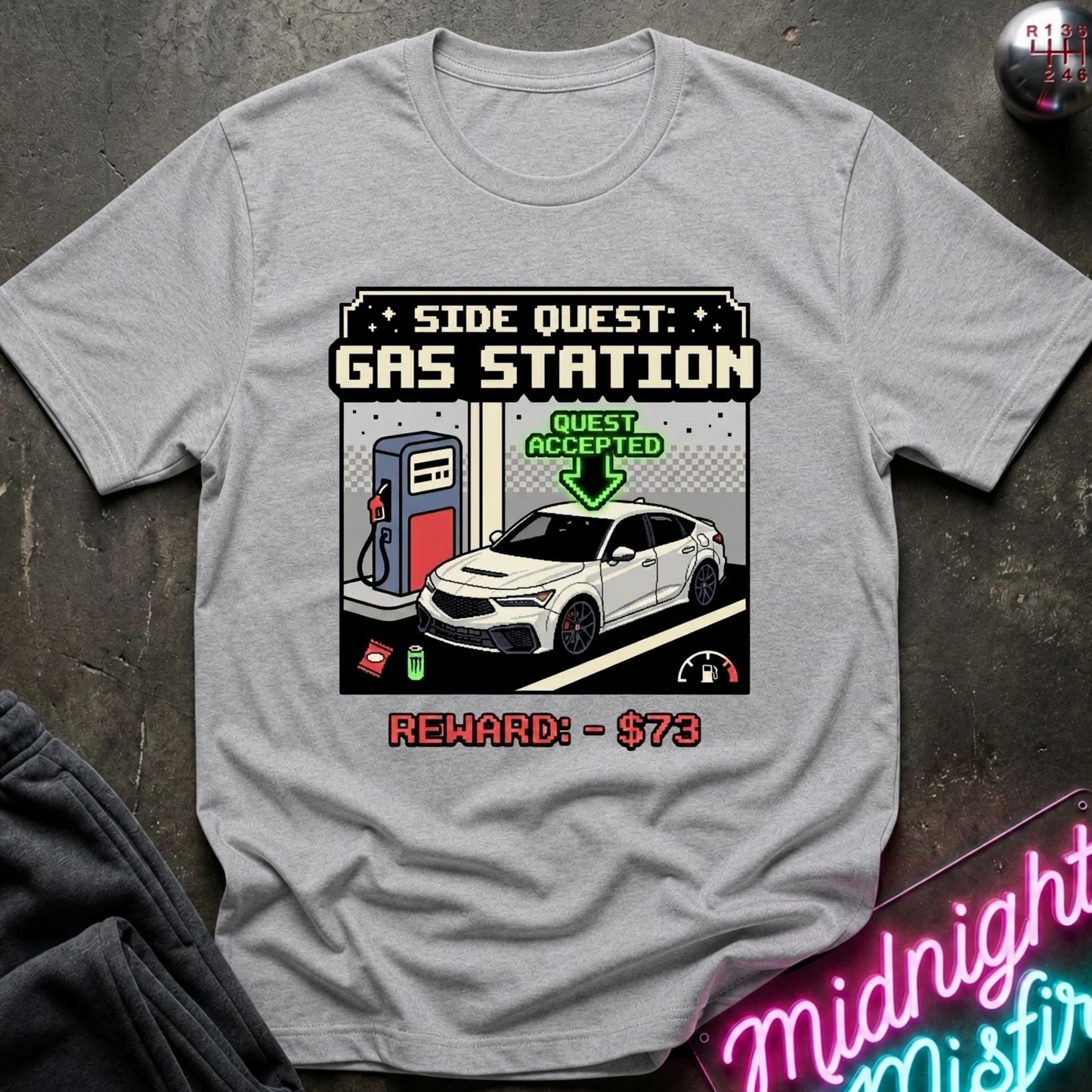 A light gray crewneck t-shirt featuring a retro pixel-art graphic of a gas station scene with a white car, a gas pump, and text reading 'SIDE QUEST GAS STATION' and 'REWARD: - $73'. The shirt is displayed flat on a dark textured surface with a neon sign and dark fabric partially visible at the edges.