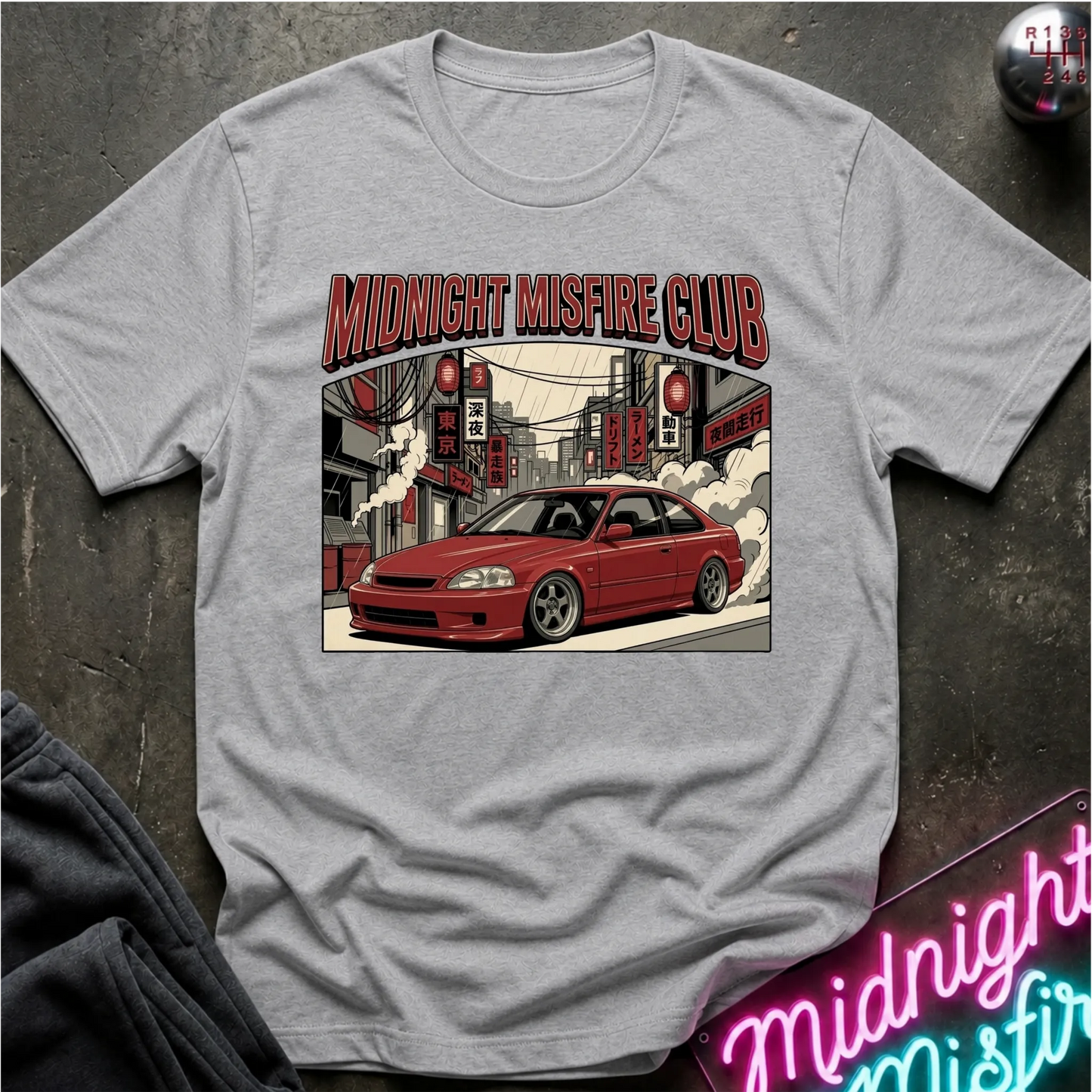 A light gray crewneck t-shirt featuring a graphic print of a red sports coupe with the text 'MIDNIGHT MISFIRE CLUB' above it, laid flat on a dark textured surface with a neon sign and sweatpants partially visible.