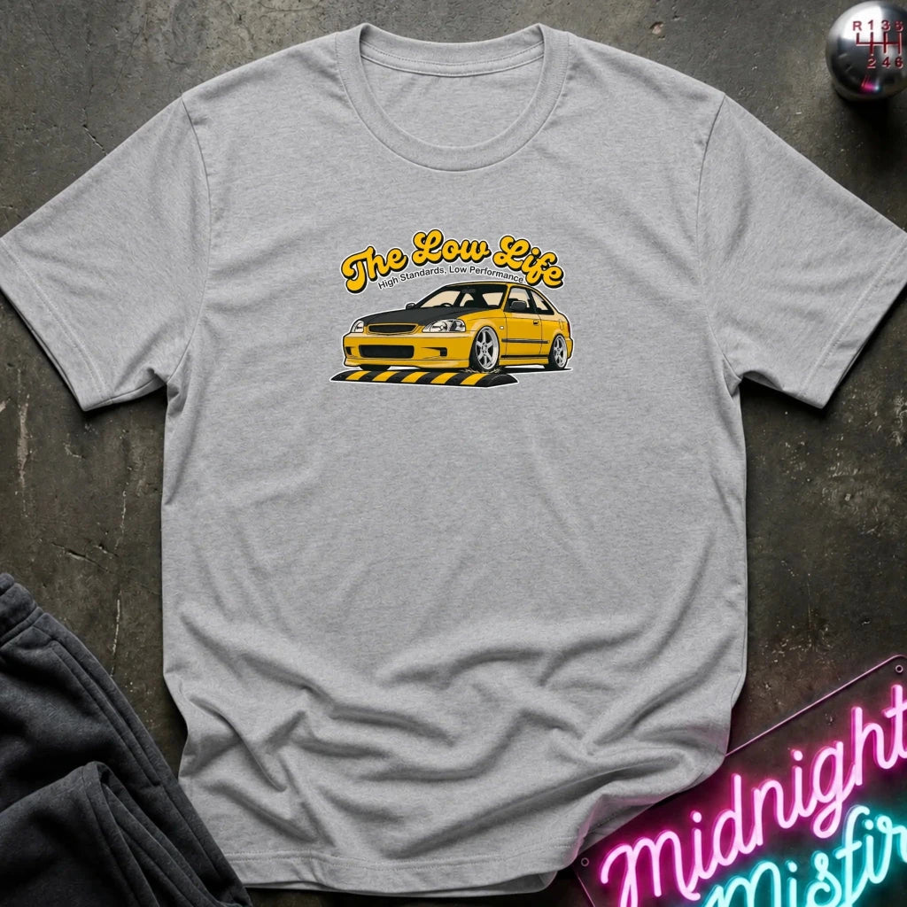 A light heather gray t-shirt laid flat on a dark textured surface, featuring a graphic of a yellow sports car with the text 'The Low Life' in yellow script. 