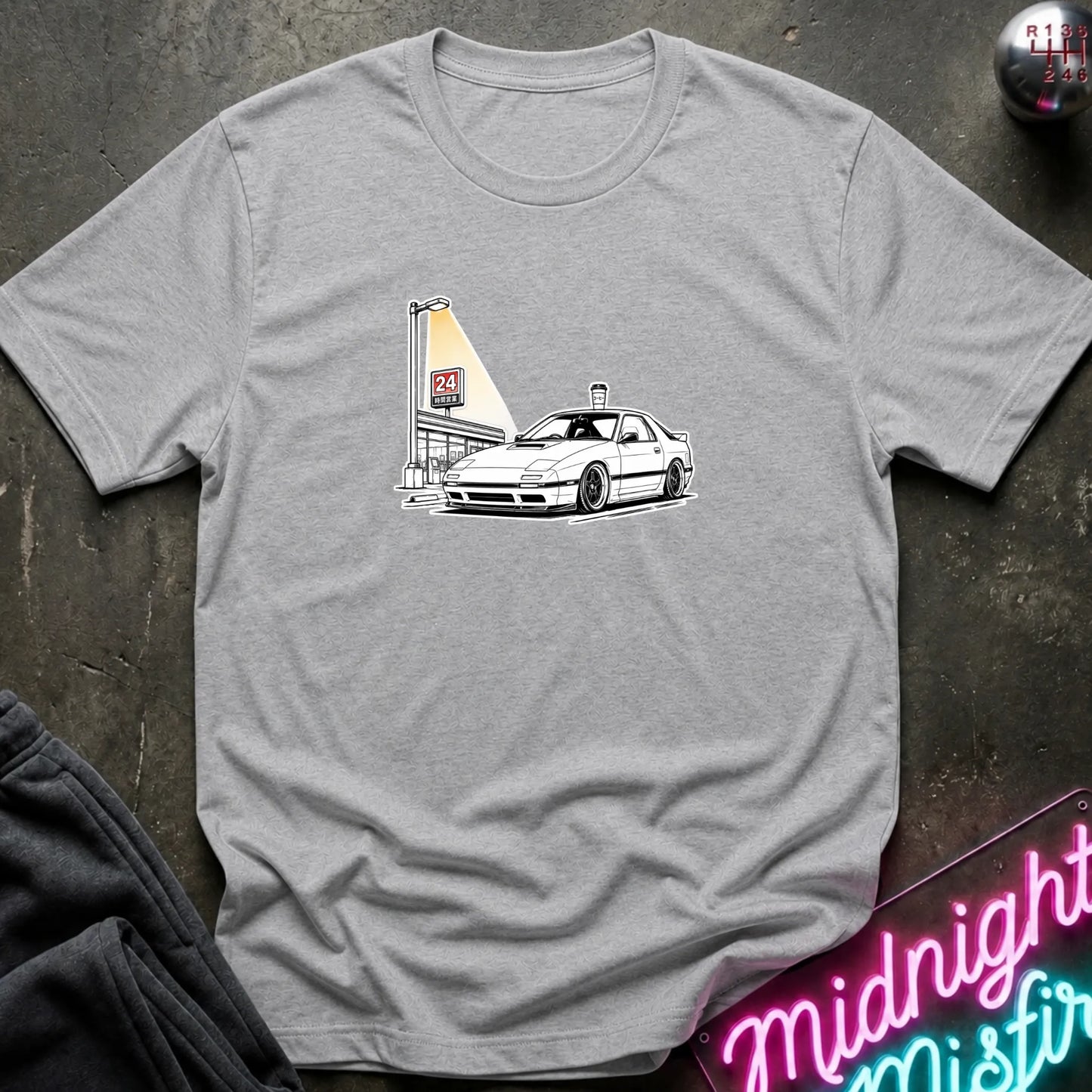 A light gray heather crewneck t-shirt featuring a graphic print of a white sports car next to a tall illuminated sign, laid flat on a dark textured surface.
