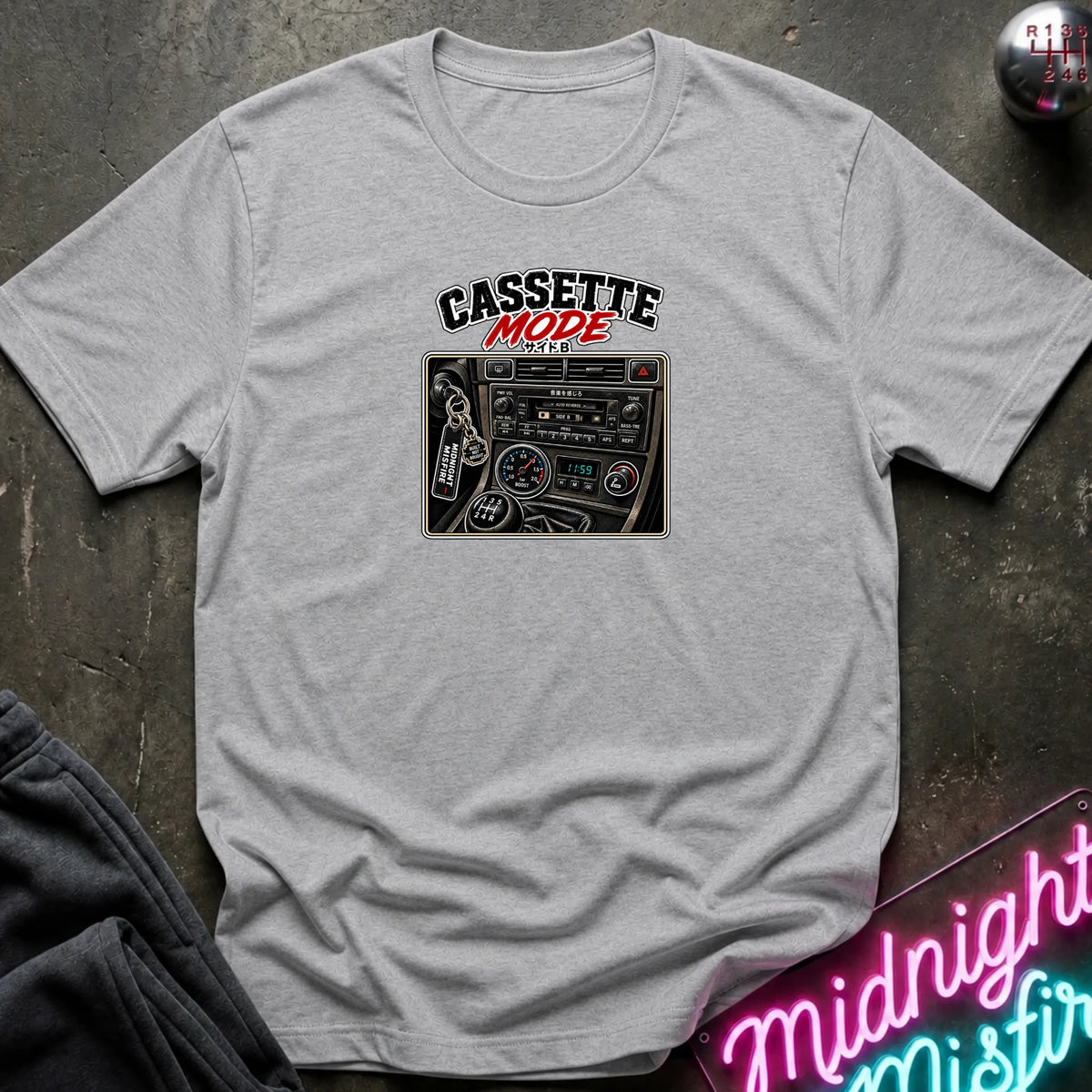 A gray short-sleeve t-shirt laid flat on a dark textured surface, featuring a graphic print of a cassette radio with the text 'CASSETTE MODE' in black and red. A neon sign and dark pants are partially visible as props.