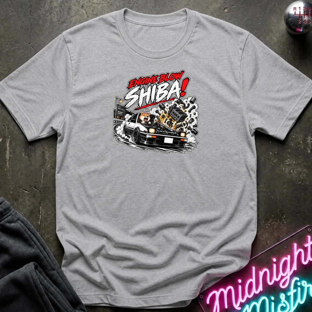 A light gray heather short-sleeve graphic t-shirt laid flat on a dark textured surface. The shirt features a stylized graphic of a white sports car with a glowing engine and the text 'ENGINE BLOW SHIBA!' in red, white, and black.