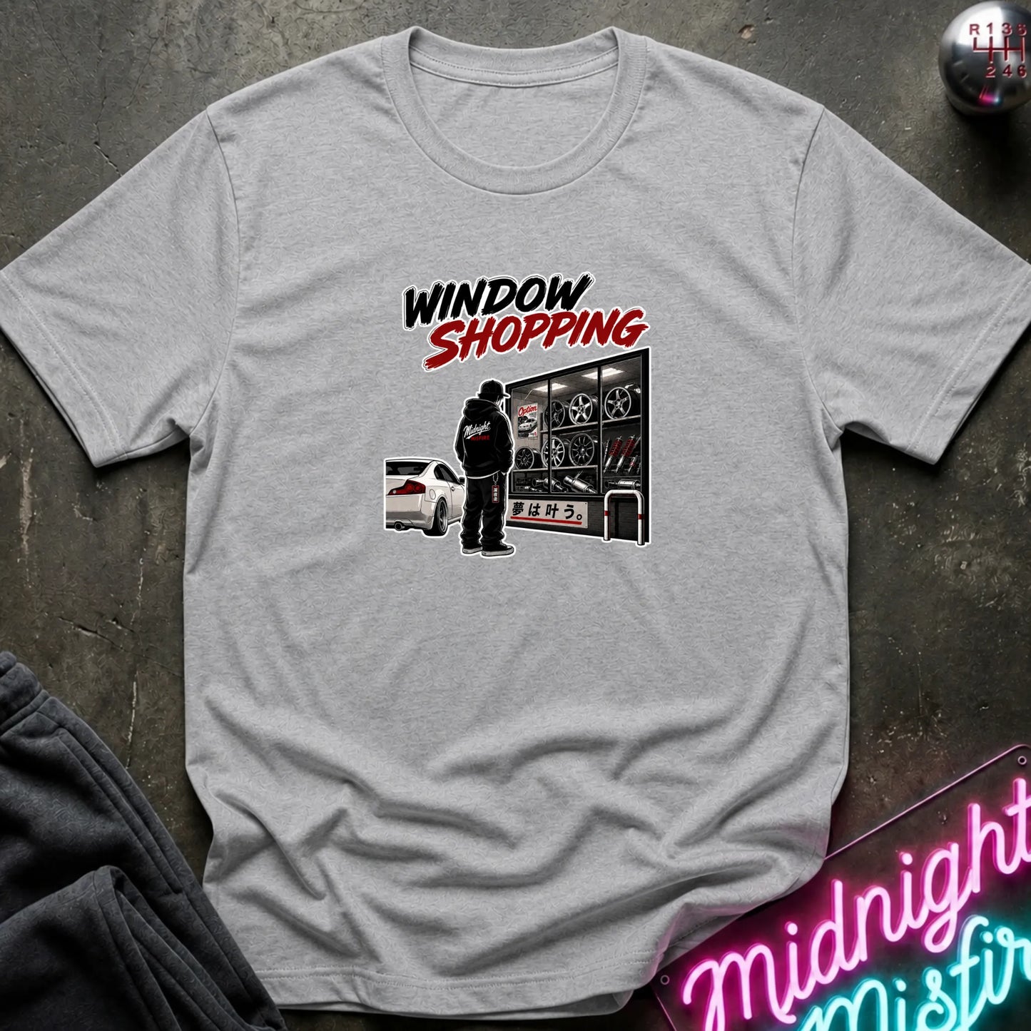 A heather gray short-sleeved t-shirt  that has a cars parked and a person checking out windows of parts.