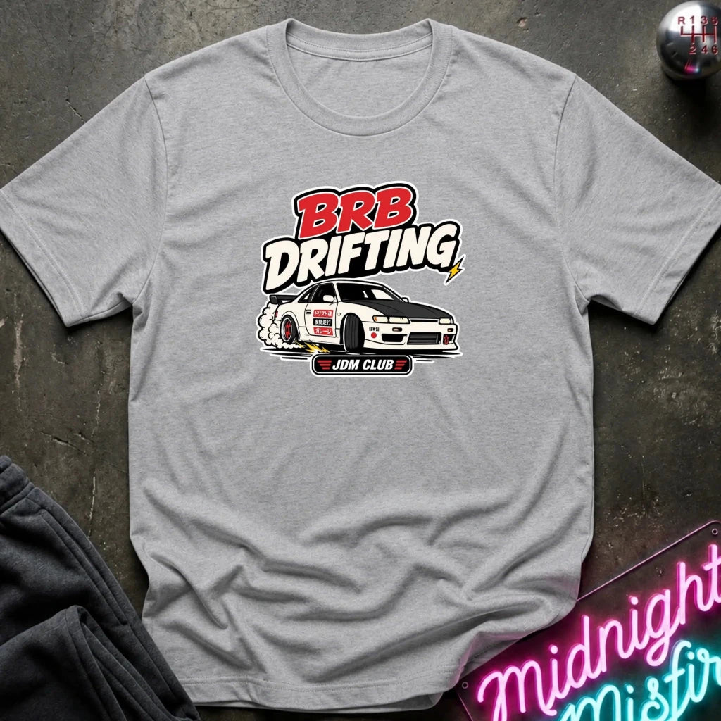 A heather gray short-sleeve t-shirt with a 'BRB DRIFTING' graphic featuring a white drift car  laid flat on a dark textured surface with a neon sign and dark pants partially visible.
