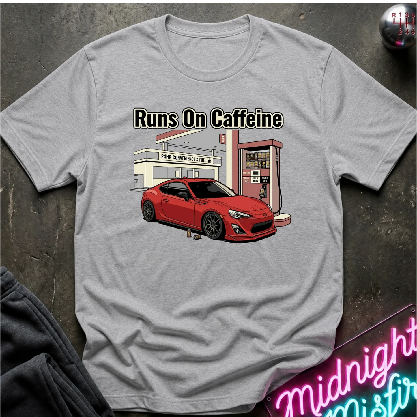 A light gray crewneck t-shirt laid flat on a dark textured surface. The shirt features a graphic print of a red sports car parked in front of a gas station, with the text 'Runs On Caffeine' printed above it.