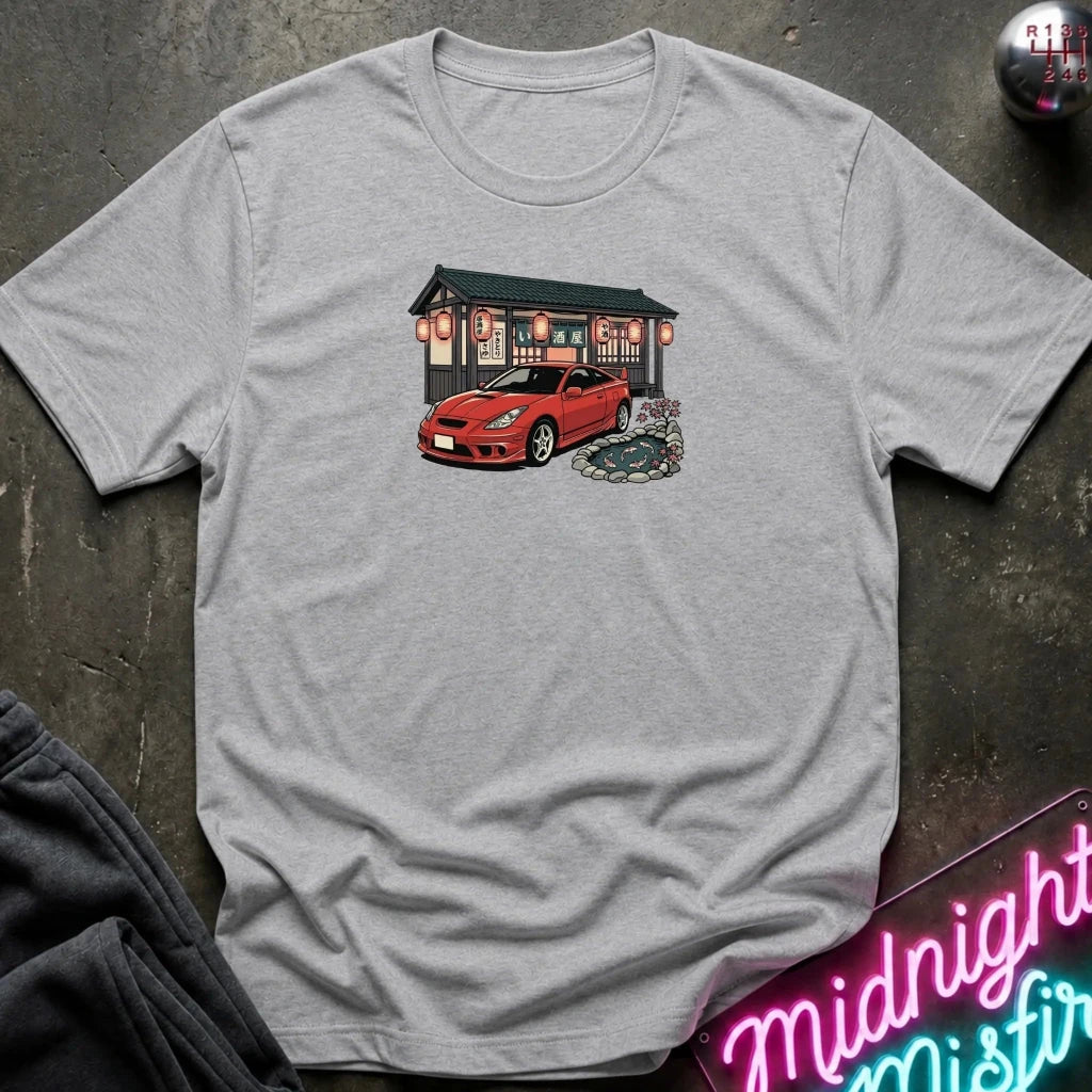 A light gray short-sleeve crewneck t-shirt laid flat on a dark textured surface, featuring a graphic print of a red sports car parked in front of a Japanese-style building with red lanterns.