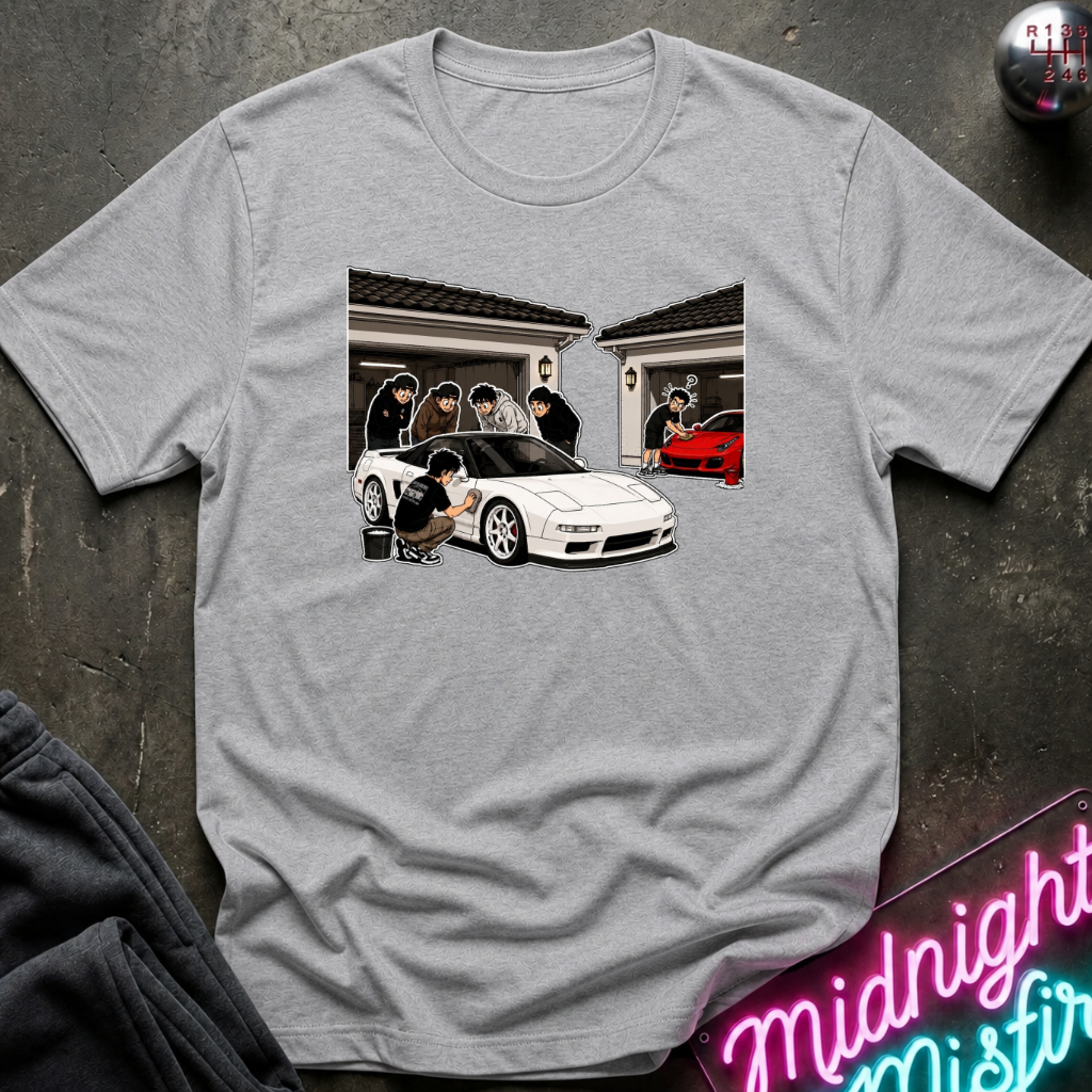 A light gray crewneck t-shirt featuring a graphic print of a white sports car and people in front of a house, laid flat on a dark textured surface with a neon sign and dark pants partially visible.