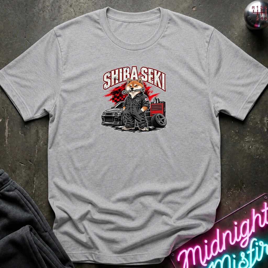 A heather gray short-sleeve t-shirt laid flat on a dark textured surface, featuring a graphic of a fox character in a black suit standing next to a black car with red and white 'SHIBA SEKI' text above.