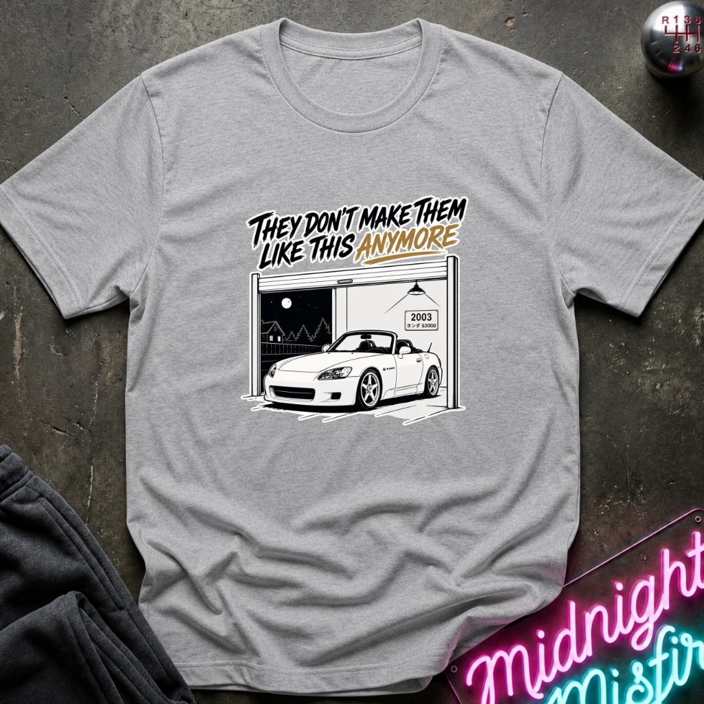 A heather gray short-sleeve t-shirt laid flat on a dark textured surface. The shirt features a graphic print of a white convertible sports car parked in front of an open garage with the text 'THEY DON'T MAKE THEM LIKE THIS ANYMORE' in black and orange lettering.
