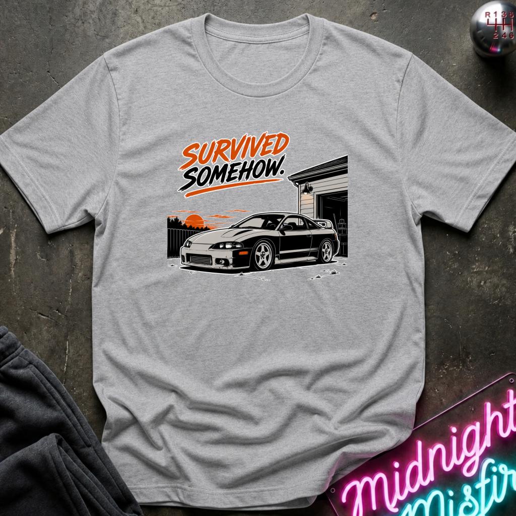 A light gray heather crewneck t-shirt featuring a graphic print of a black sports car in front of a garage with the text 'SURVIVED SOMEHOW.' in orange and black.