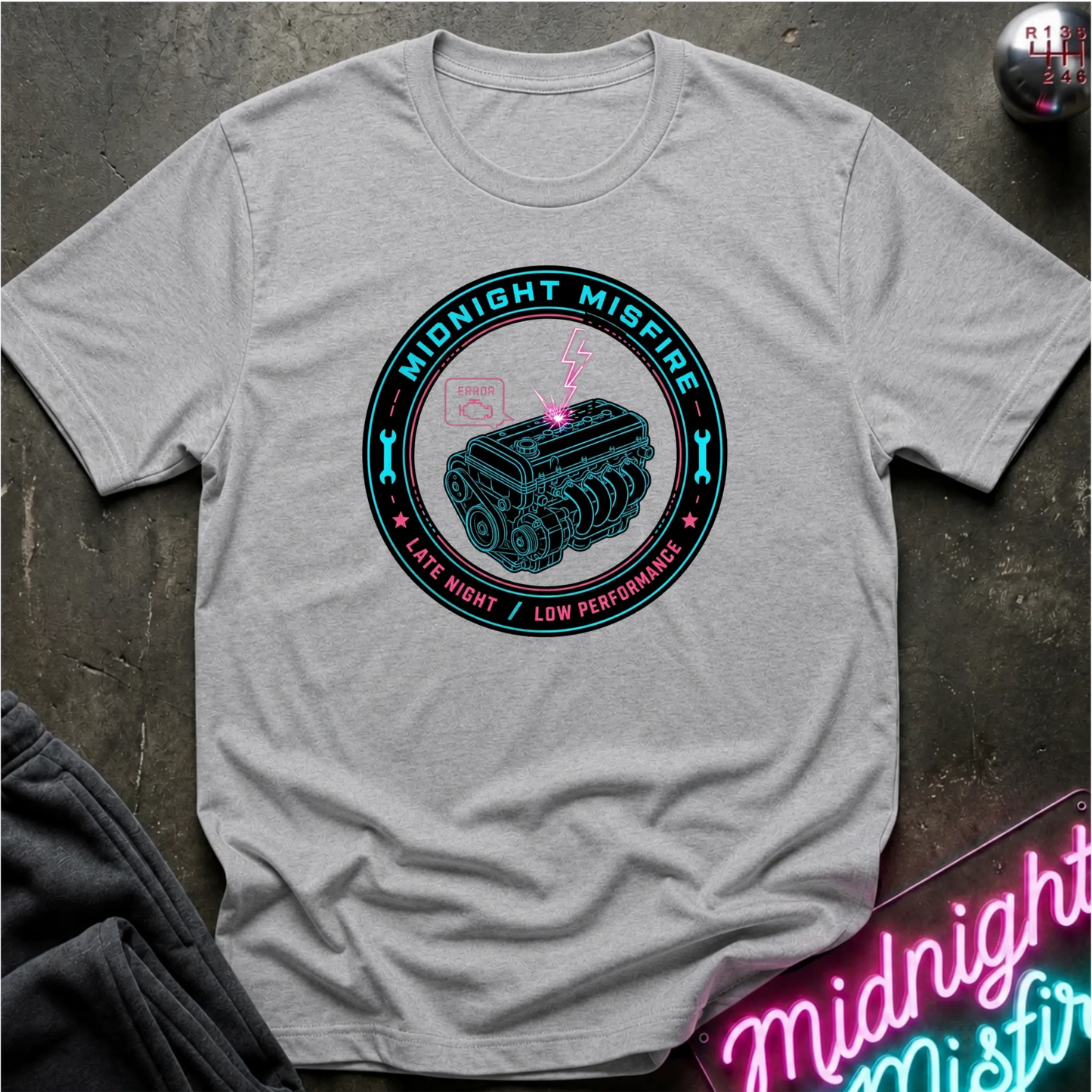 A light gray heather crewneck t-shirt laid flat on a dark textured surface. The shirt features a circular graphic in the center with a teal engine illustration, a pink lightning bolt, and teal and pink text reading 'MIDNIGHT MISFIRE' and 'LATE NIGHT / LOW PERFORMANCE'.