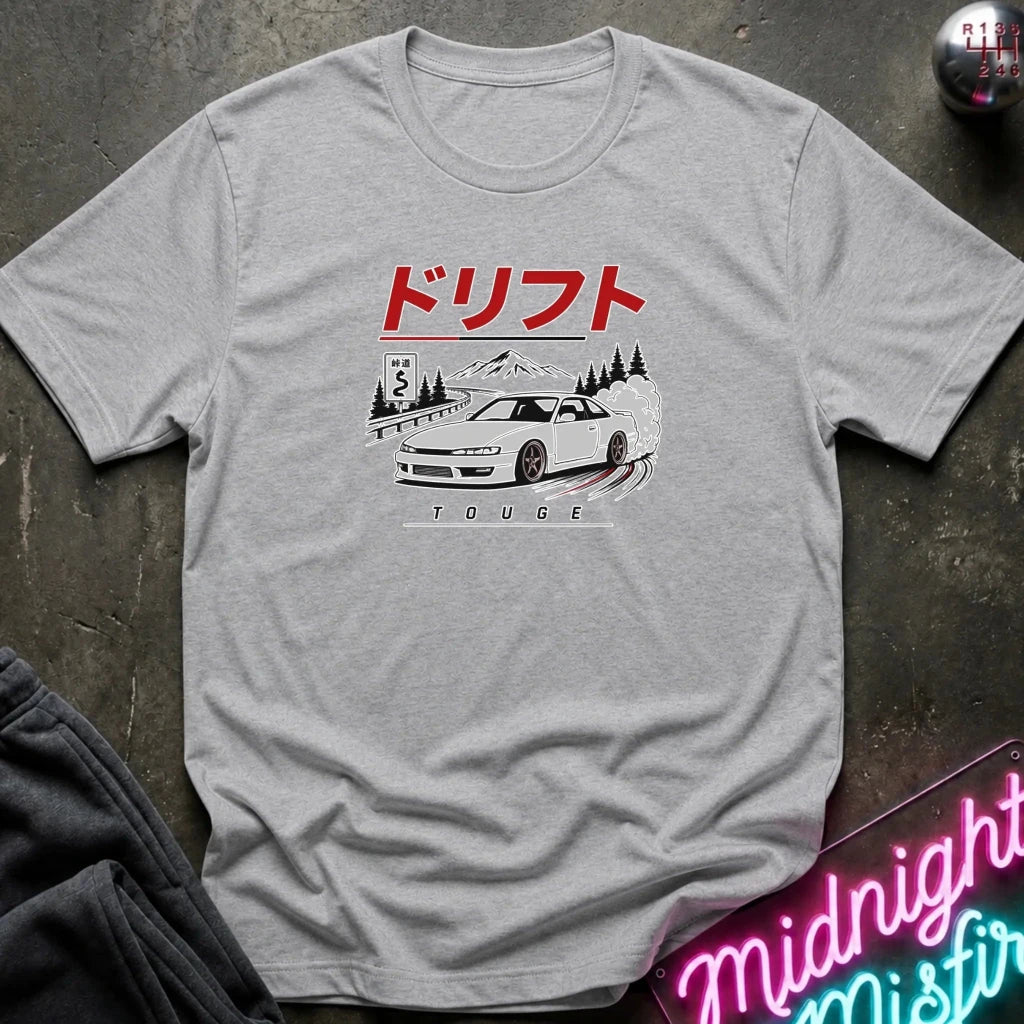 A light gray heather crewneck t-shirt featuring a graphic print of a white sports car drifting on a mountain road, with red Japanese text above and 'TOUGHE' below. The shirt is laid flat on a dark textured surface with a neon sign and dark pants partially visible.