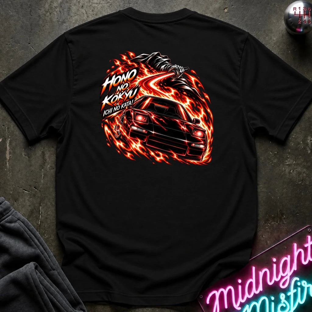 black short-sleeve t-shirt laid flat on a dark textured surface, showing the back graphic with a car and the text "ichi no kata!"