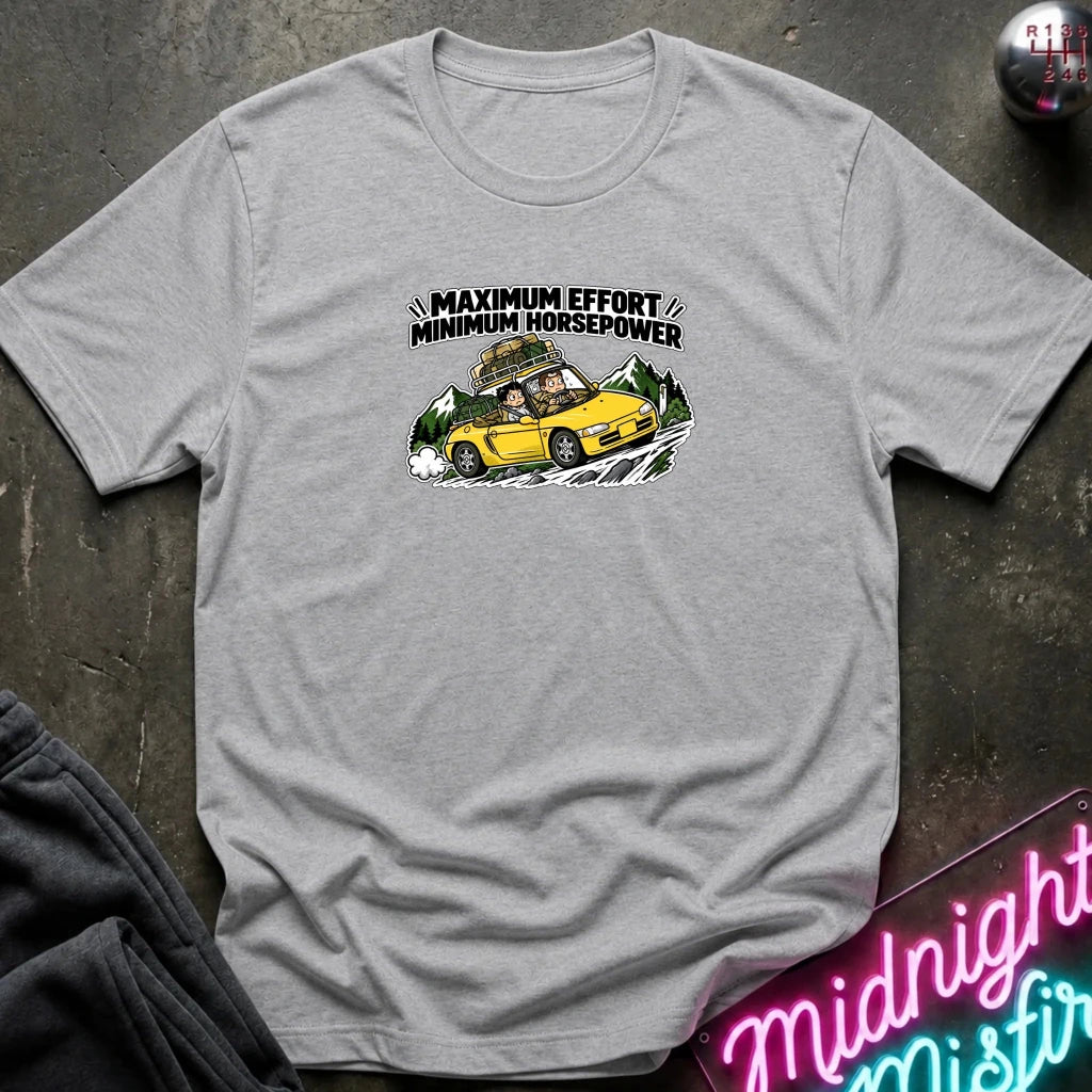 gray graphic t-shirt laid flat on a dark textured surface. The graphic features a yellow sports car with a driver and passengers, with the text 'MAXIMUM EFFORT MINIMUM HORSEPOWER' above it.