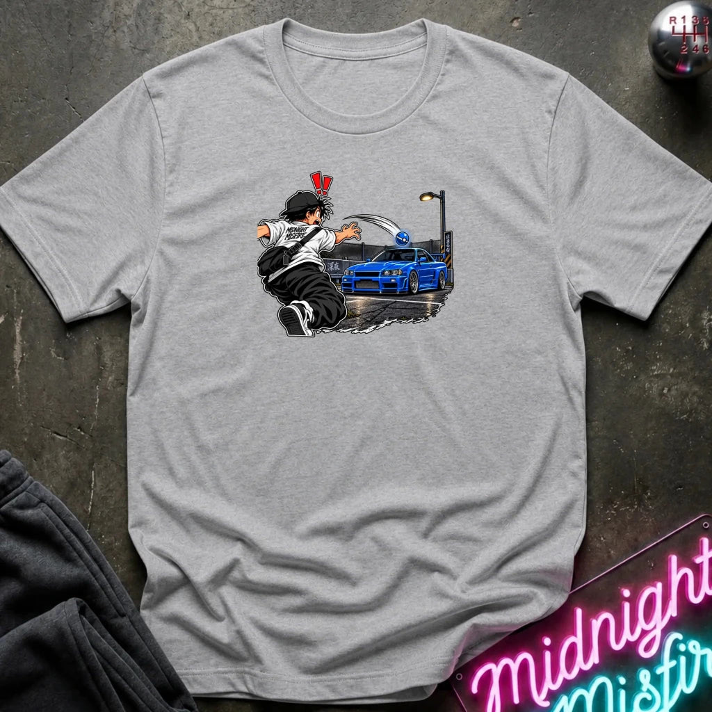 A light heather gray short-sleeve crewneck t-shirt laid flat on a dark textured surface. The center of the shirt features a graphic print of an anime character in a black and white outfit with a hat, next to a blue sports car under a streetlamp.