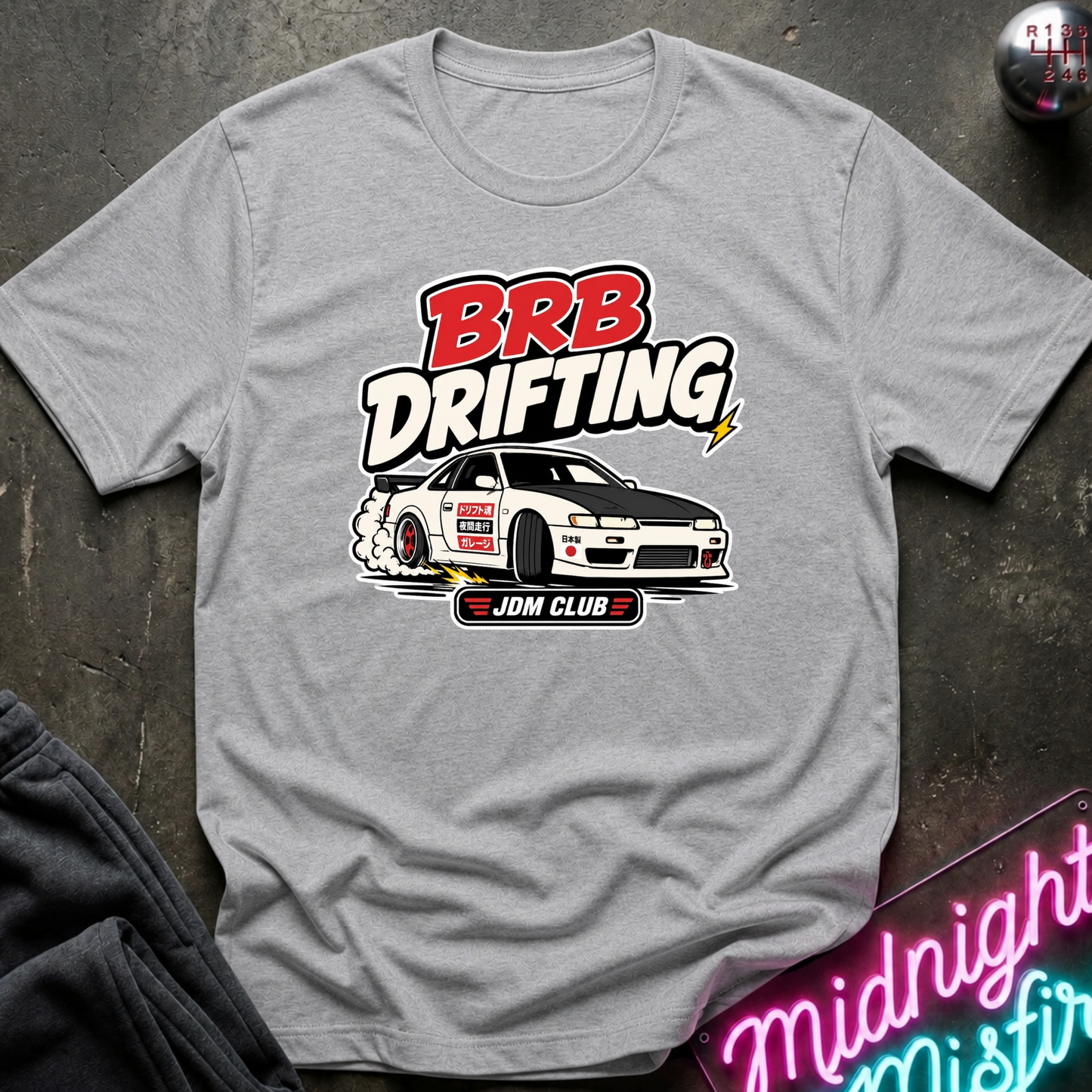 A heather gray short-sleeve t-shirt with a 'BRB Drifting' graphic featuring a white and black drifting car, laid flat on a dark textured surface with a neon sign and dark pants partially visible.