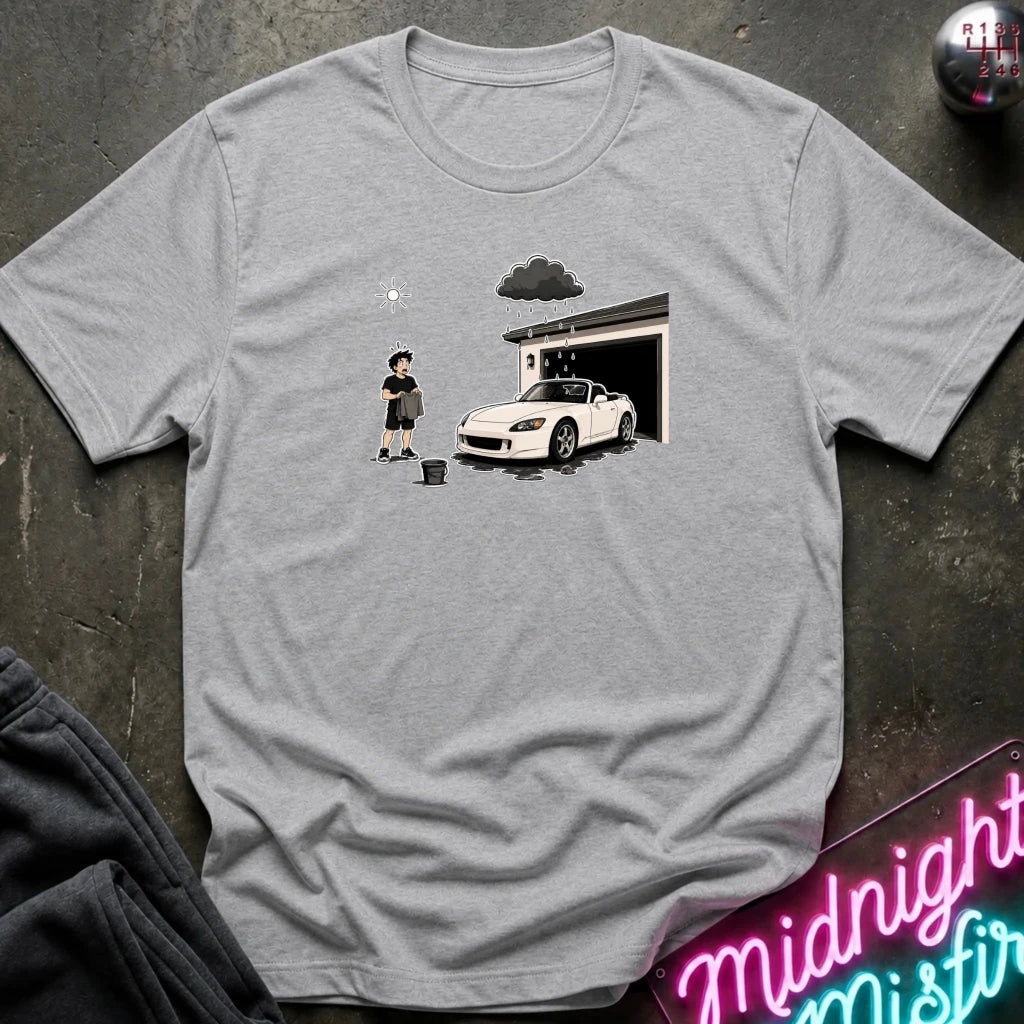 A light gray heather crewneck t-shirt laid flat on a dark textured surface. The shirt features a graphic print of a cartoon character washing a white sports car in front of a garage under a dark cloud.
